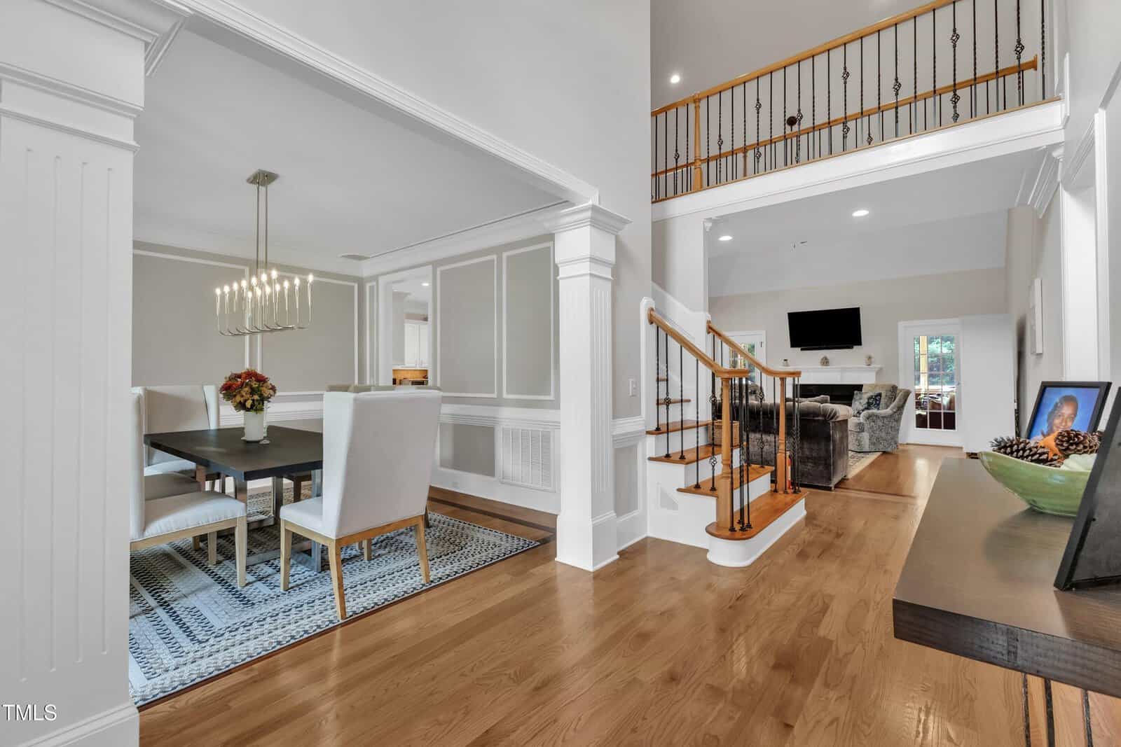 A view of a home interior shows a dining area with a dark table and upholstered chairs on the left, a staircase with wooden steps and black railings, and a living room with a TV and sofas in the background.