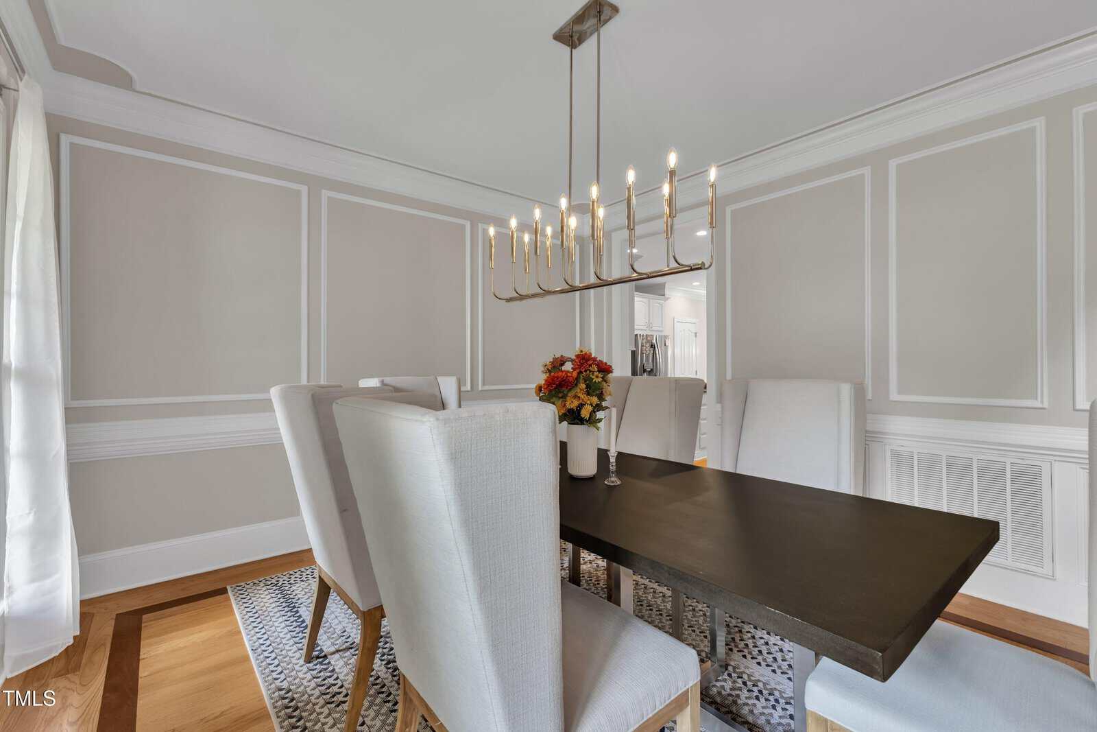 A dining room with a rectangular dark wood table, six high-back upholstered chairs, a modern rectangular chandelier, light-colored walls with decorative molding, and a vase of flowers on the table.