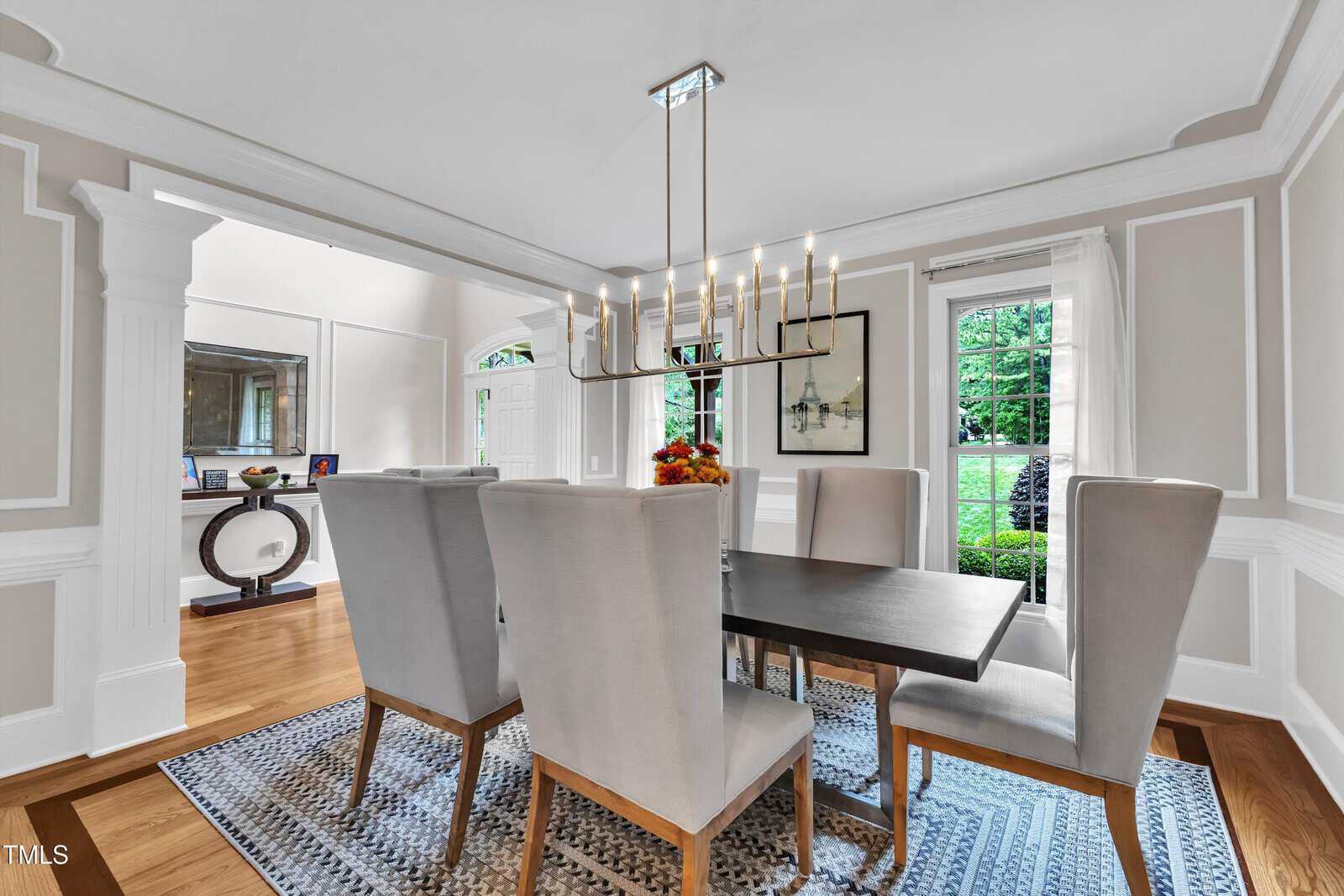 A dining room with a rectangular table, six high-back chairs, a modern chandelier, a wall mirror, artwork, large windows, and hardwood floors with a patterned rug.