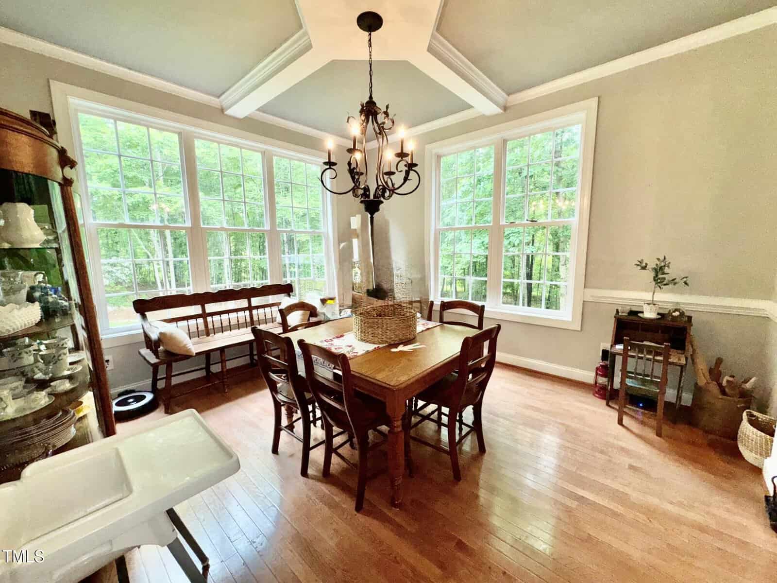 A dining room with hardwood floors, a wooden table and six chairs, a bench by large windows, a chandelier, and various decorative items and furniture along the walls. Trees are visible through the windows.