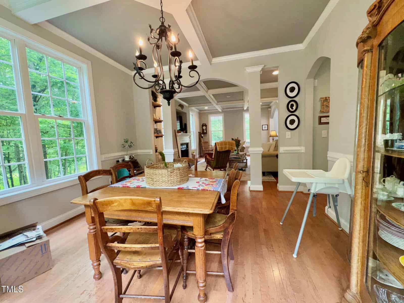 A dining area with a wooden table and chairs sits near large windows; a black chandelier hangs above. In the background, an open living room and kitchen are visible. A high chair is placed near a glass cabinet.