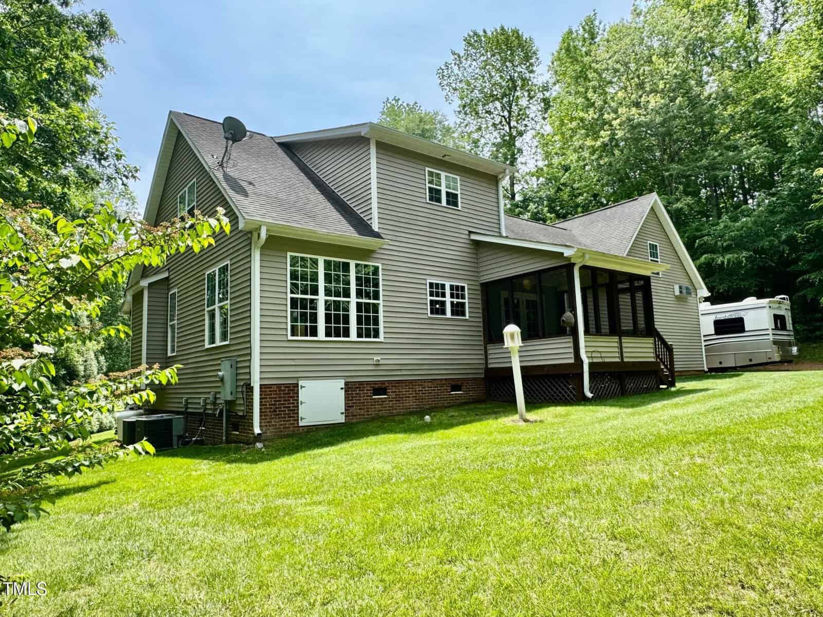 A two-story suburban house with beige siding, white trim, and a screened porch sits on a grassy lawn surrounded by trees. A white RV is parked in the background near the house.