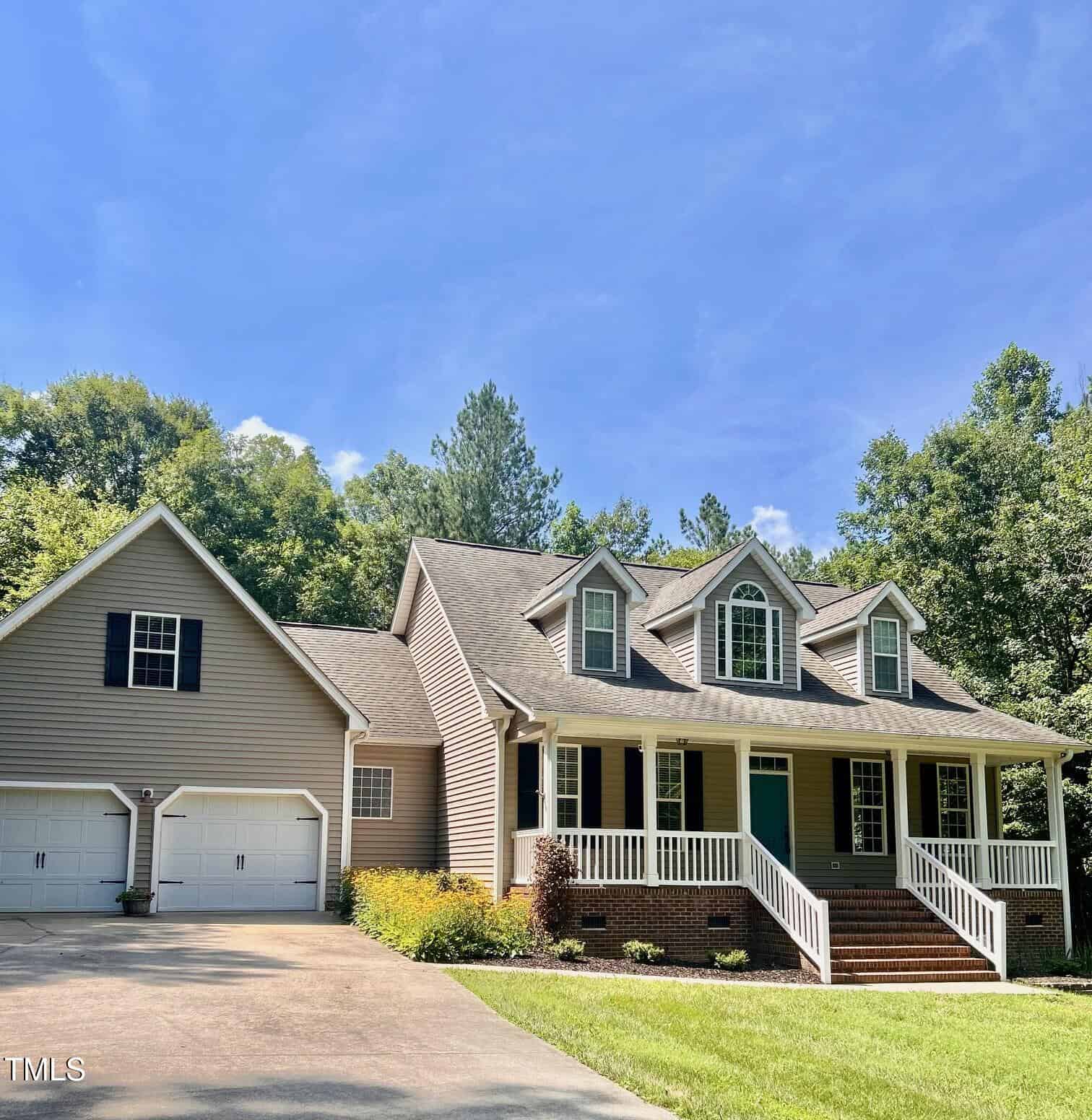 A tan, two-story house with a front porch, three dormer windows, white trim, and a two-car garage sits on a sunny day, surrounded by green trees and a well-kept lawn with yellow flowers by the driveway.