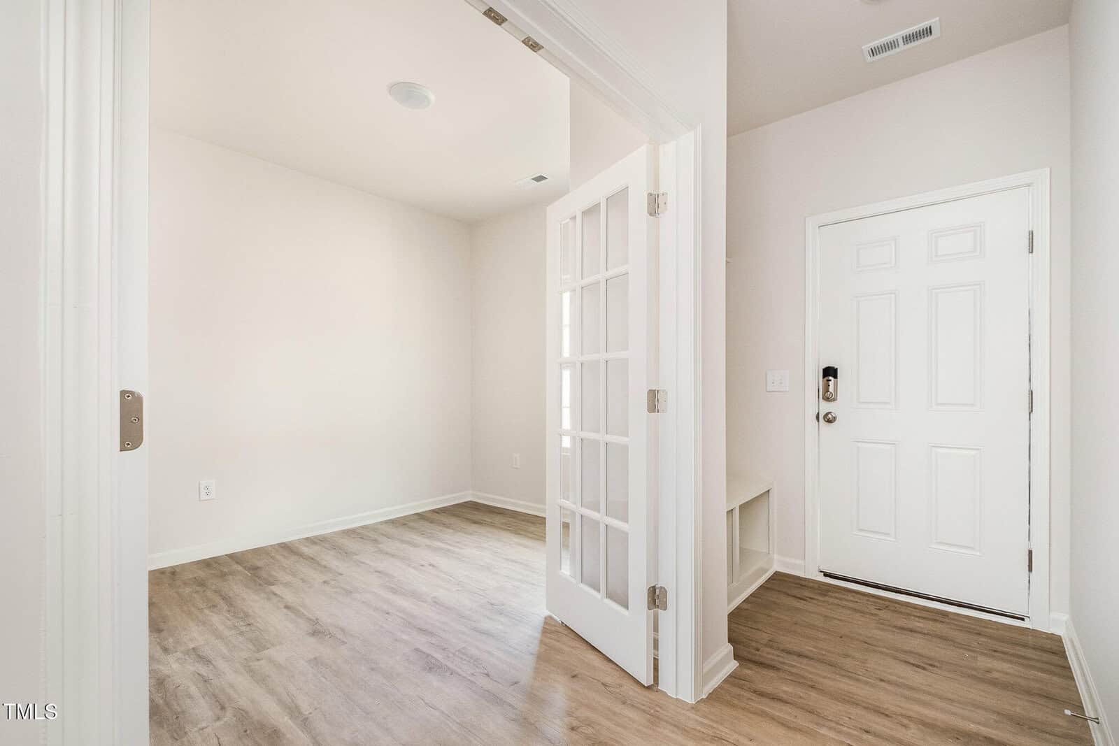 A small, empty room with light-colored walls and wood-style flooring, viewed through glass French doors near a white front door. The space is well-lit with natural light.