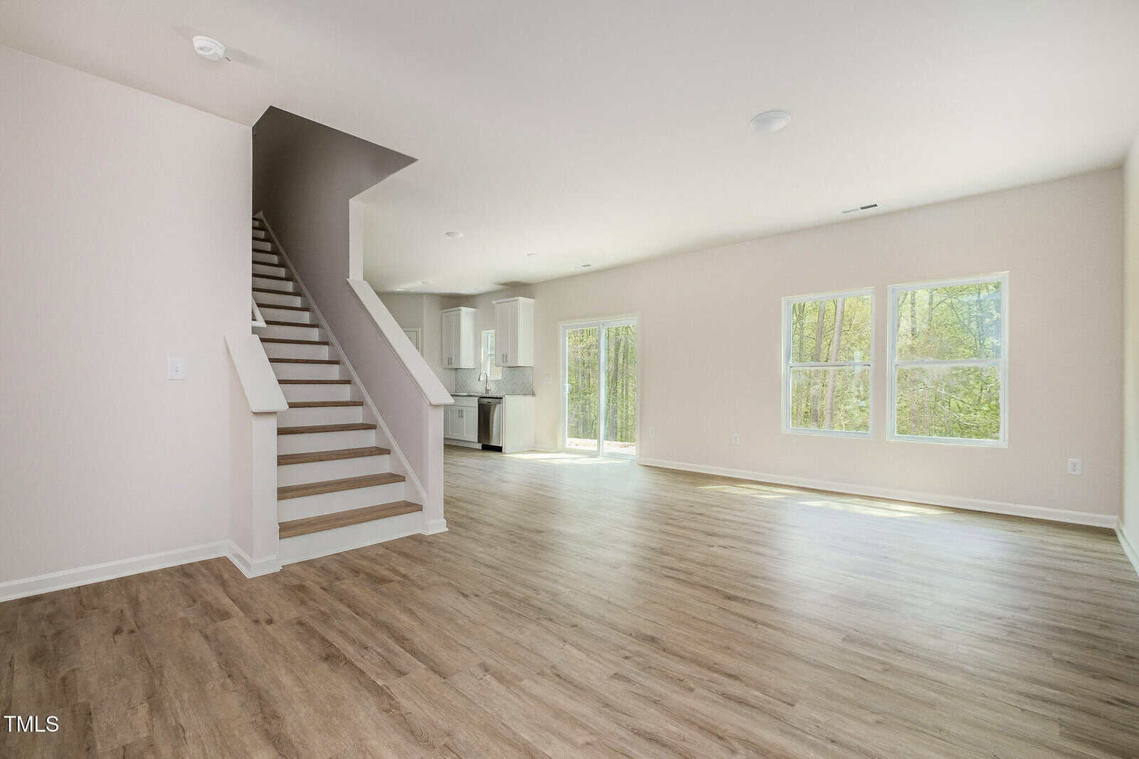 A spacious, empty room with light-colored walls and wood flooring, featuring a staircase on the left, large windows, and an open view into a modern kitchen area. Natural light fills the space.