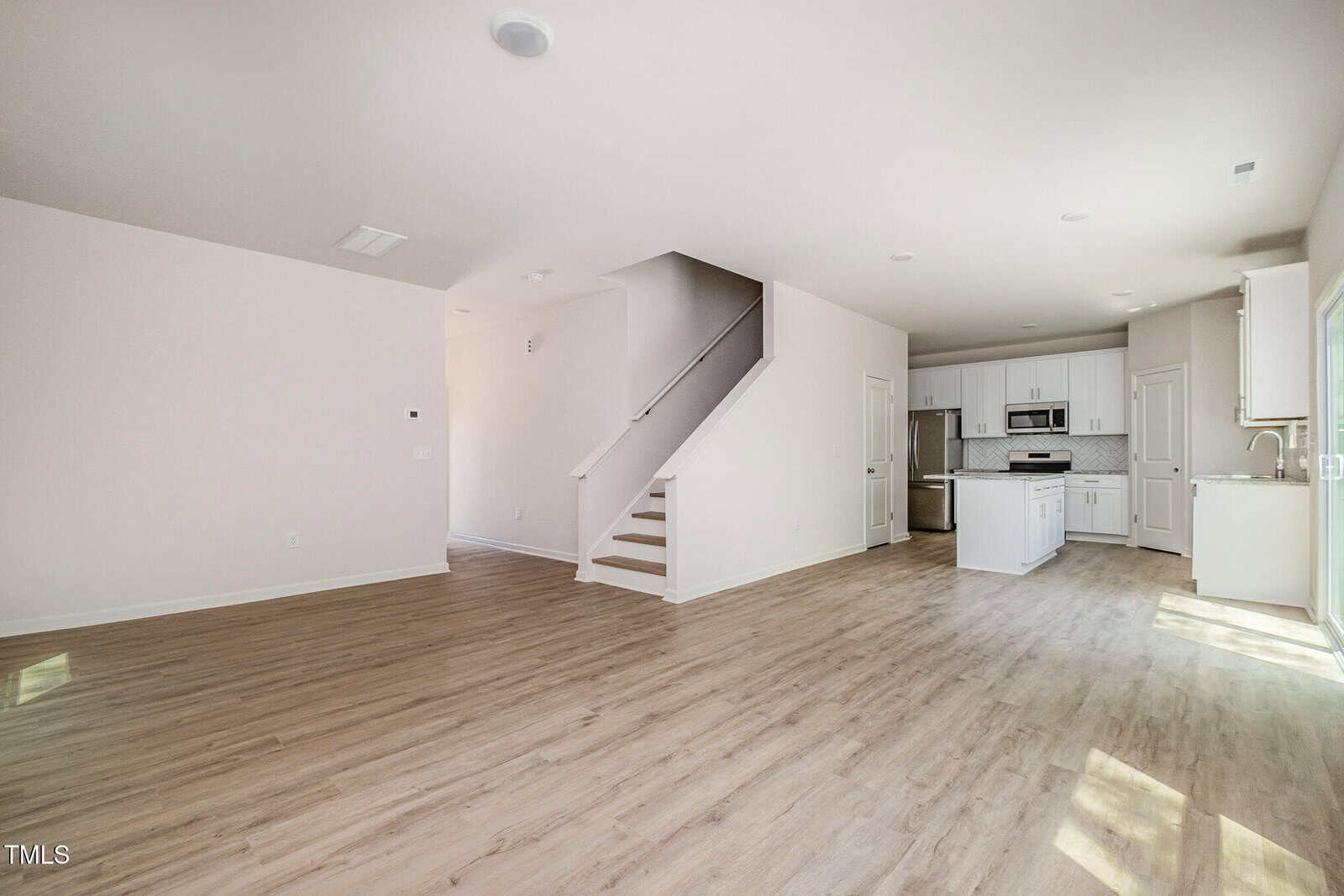 Open-concept living space with light wood flooring, white walls, and a staircase leading upstairs. The kitchen features white cabinets, stainless steel appliances, and an island. Natural light enters through several windows.