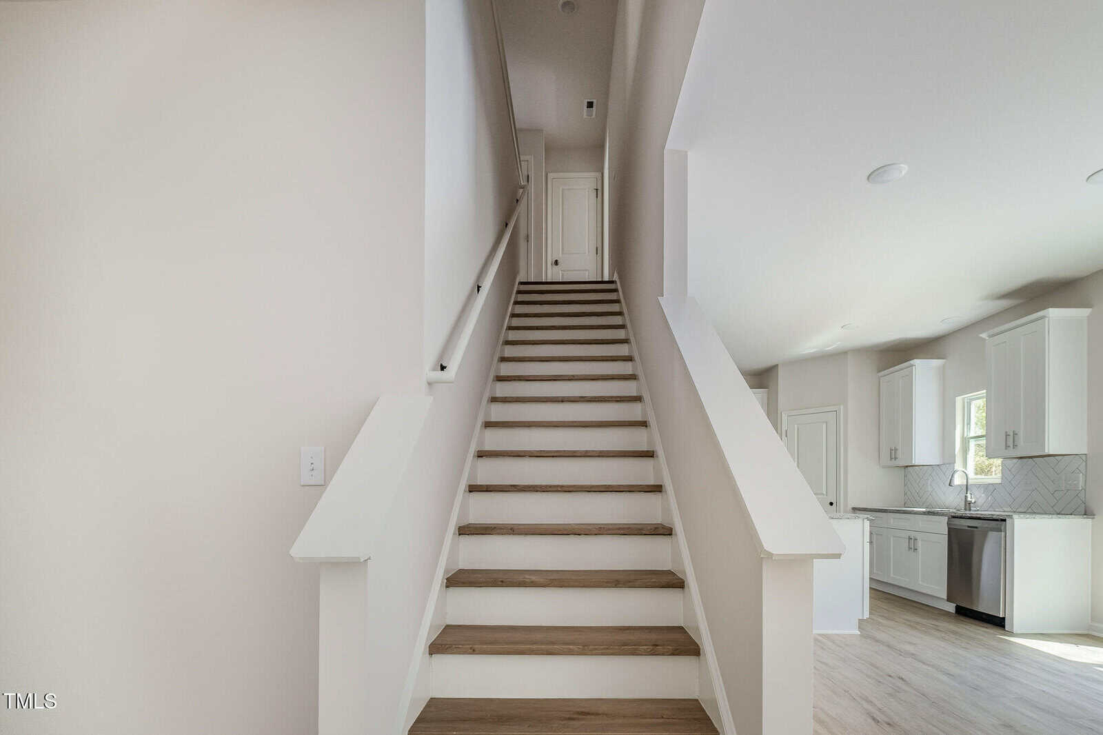 A straight staircase with light wood steps and white railings leads up to a closed door. The adjacent kitchen features white cabinets, a window, and light flooring. The walls are painted a light, neutral color.