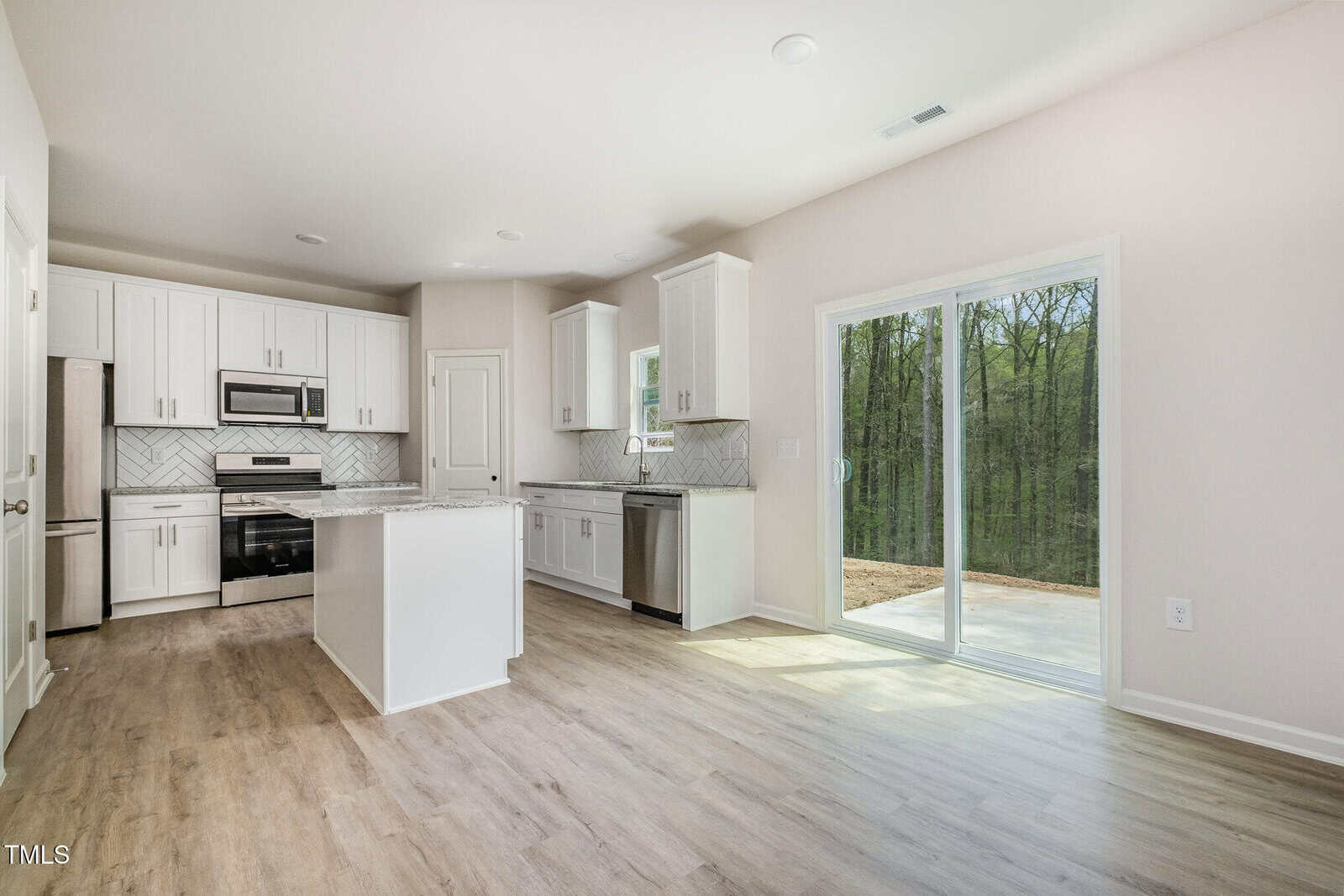 Modern kitchen with white cabinets, stainless steel appliances, a center island, and light wood flooring. Large sliding glass doors open to a patio with a forest view. The space is bright with natural light.