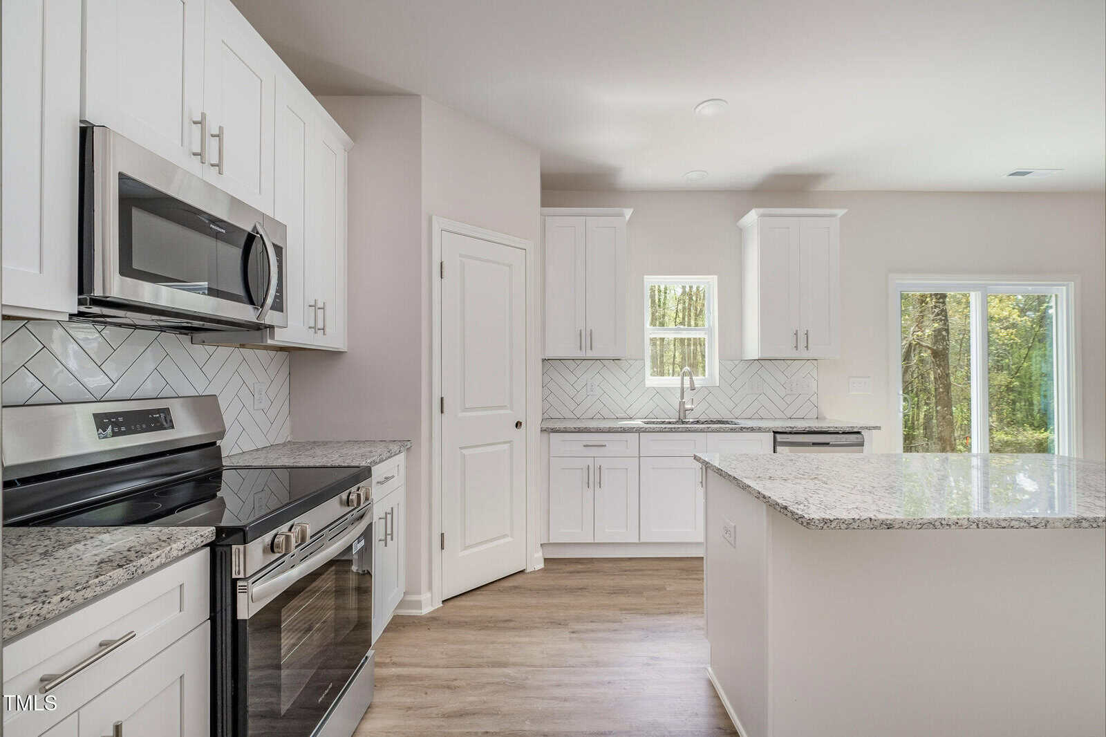 A modern kitchen with white cabinets, stainless steel appliances, a light gray herringbone tile backsplash, granite countertops, light wood flooring, and large windows letting in natural light.