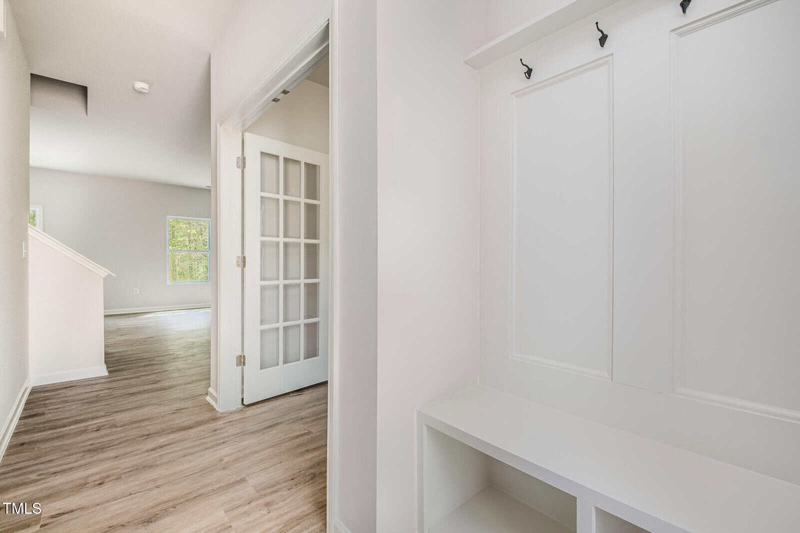 A hallway with light wood flooring, white walls, a built-in bench with hooks, a staircase, and a room with French doors and windows visible in the background.