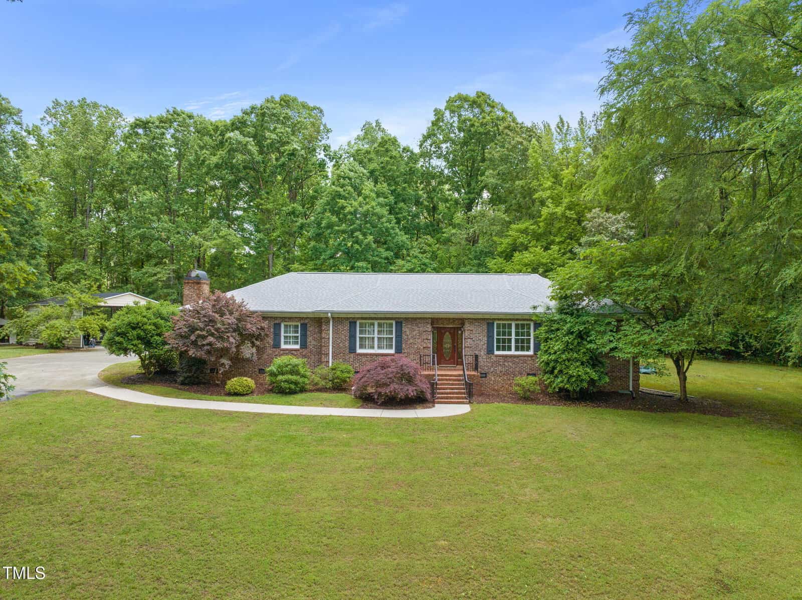 A single-story brick house with a gray roof sits on a large, neatly maintained lawn. A curved driveway leads to the front door, and the property is surrounded by mature trees and landscaping.