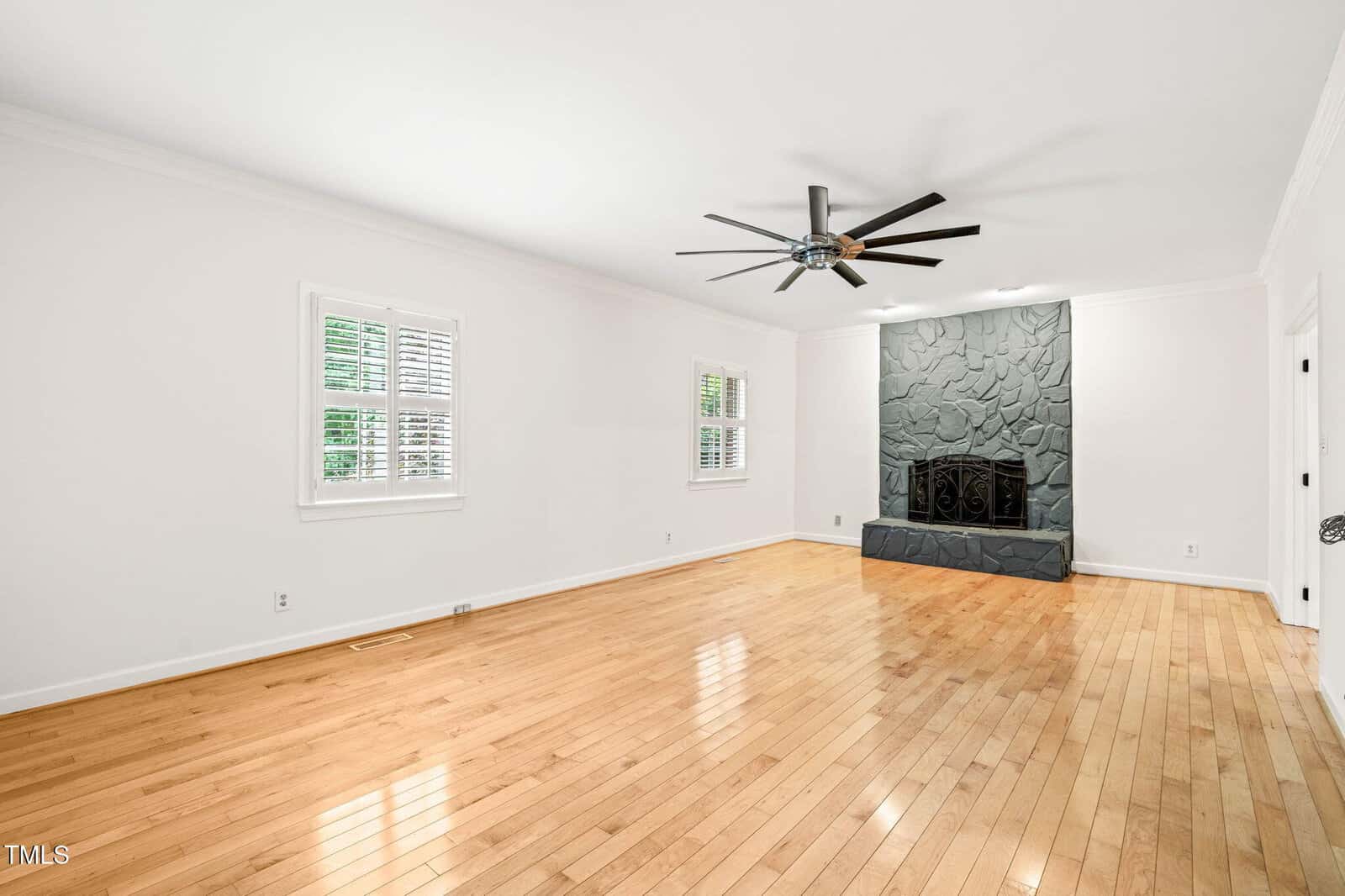 A spacious living room with light wood flooring, white walls, two windows with white shutters, and a dark stone fireplace with a large black ceiling fan overhead.