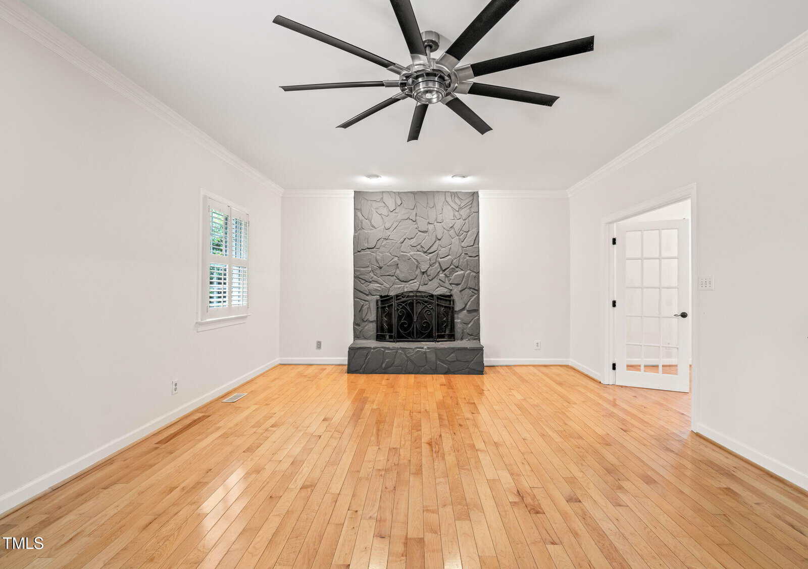 A spacious, empty living room with light wood floors, white walls, a large black ceiling fan, a stone fireplace, and doors with glass panels. One window with white shutters is on the left wall.