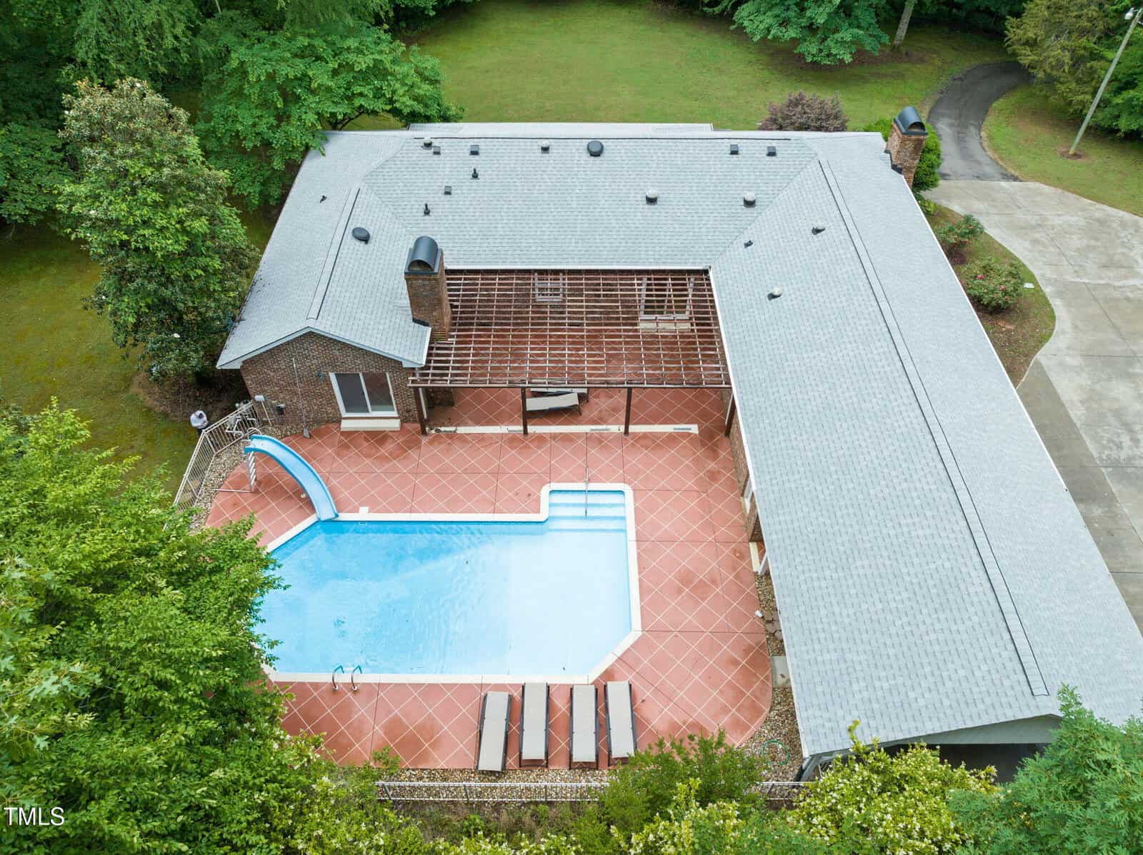 Aerial view of a large house with a gray roof at 1514 Lawrence Road in Creedmoor, surrounding an outdoor rectangular swimming pool with a slide, set on a tiled patio and bordered by lush trees and lawns.
