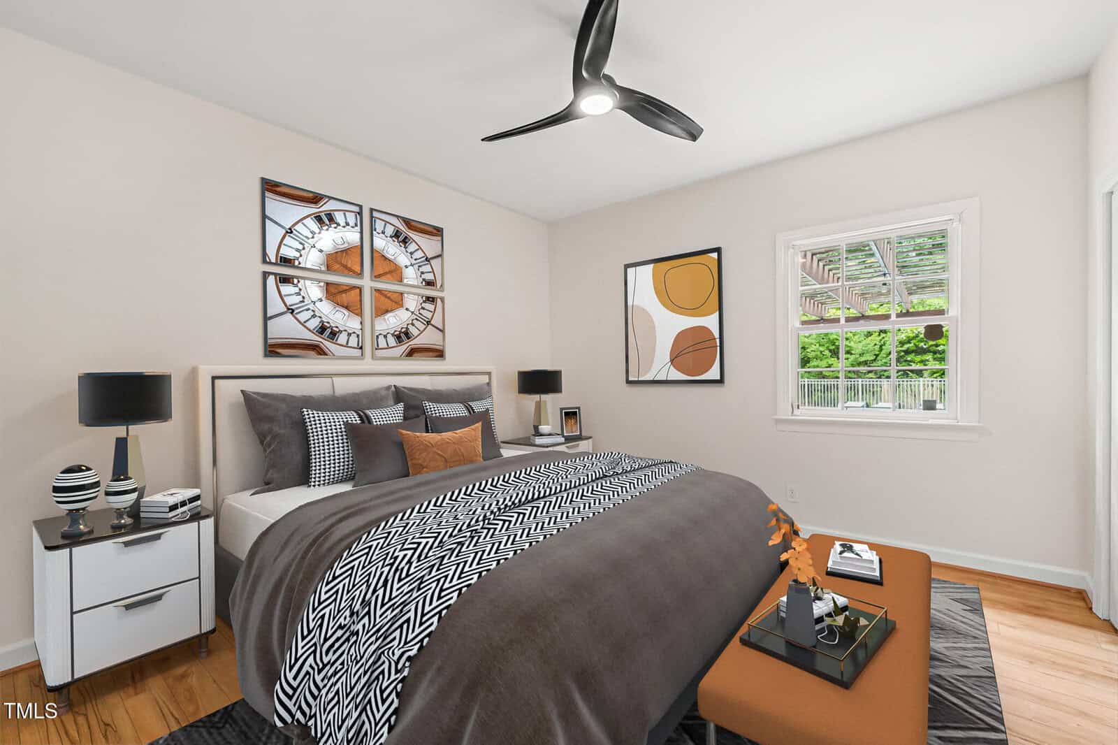 A modern bedroom with a double bed featuring gray and white bedding, two white nightstands with lamps, a brown ottoman, wall art, a ceiling fan, and a window showing trees and a fenced yard outside.