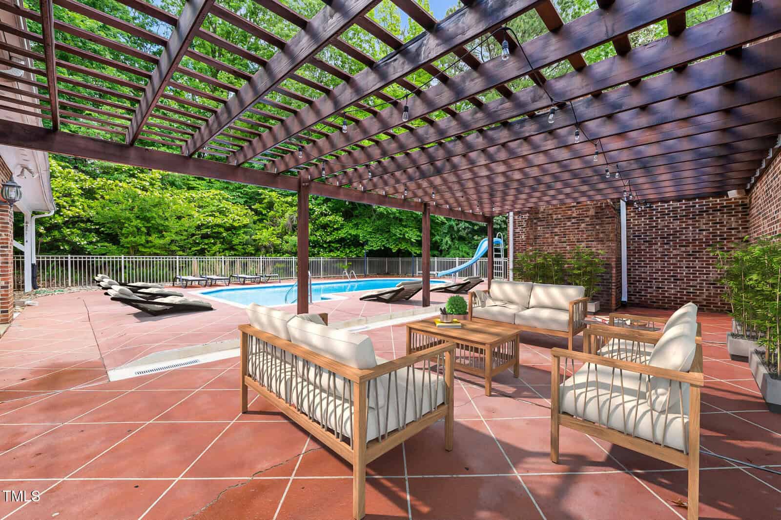Outdoor patio with a wooden pergola, cushioned seating area, potted plants, and a table, overlooking a swimming pool with lounge chairs, a railing, and a slide, surrounded by trees and a brick wall.