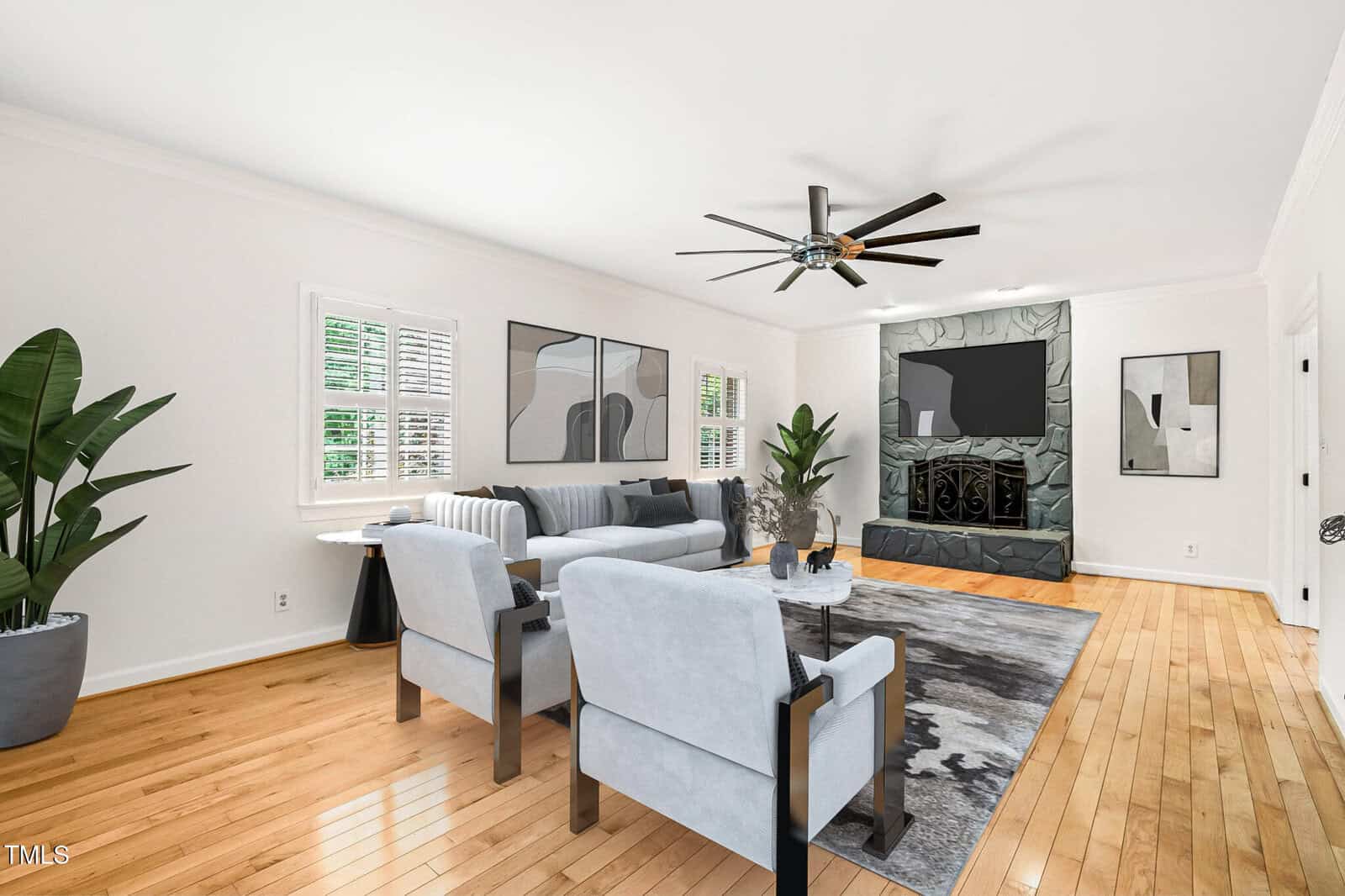 A modern living room with light hardwood floors, white walls, gray furniture, abstract wall art, a large television, two potted plants, and a ceiling fan. Natural light enters through shuttered windows.