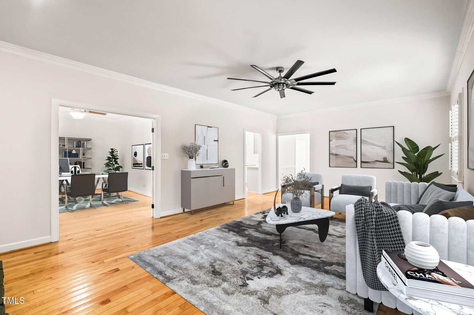 A modern living room with light walls, a ceiling fan, gray furniture, a patterned rug, and minimalist decor. An adjoining room with hardwood floors and a large plant is visible through an open doorway.