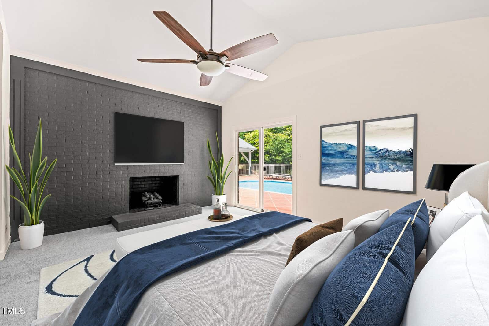 A modern bedroom with a large bed, blue and white bedding, two wall paintings, a black accent wall with a TV and fireplace, plants, and sliding glass doors opening to a pool area.