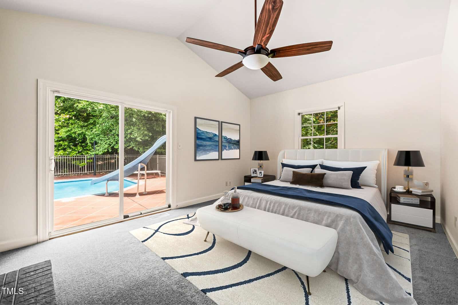 A bedroom with a bed, white bench, two nightstands, wall art, and a ceiling fan. A large window and sliding glass door open to a patio with a swimming pool and slide. The room has light walls and a patterned rug.