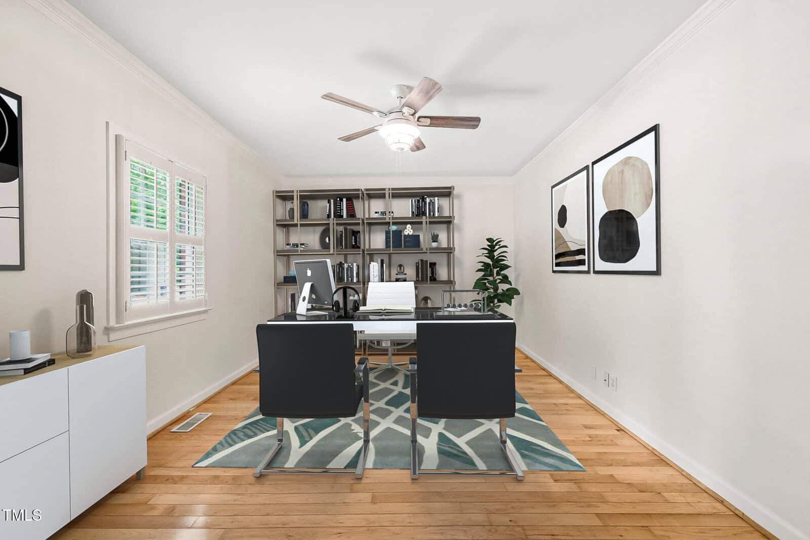 A home office with two black chairs facing a desk, a white chair behind the desk, bookshelves against the back wall, abstract art on the walls, a ceiling fan, a potted plant, and a window with white shutters.