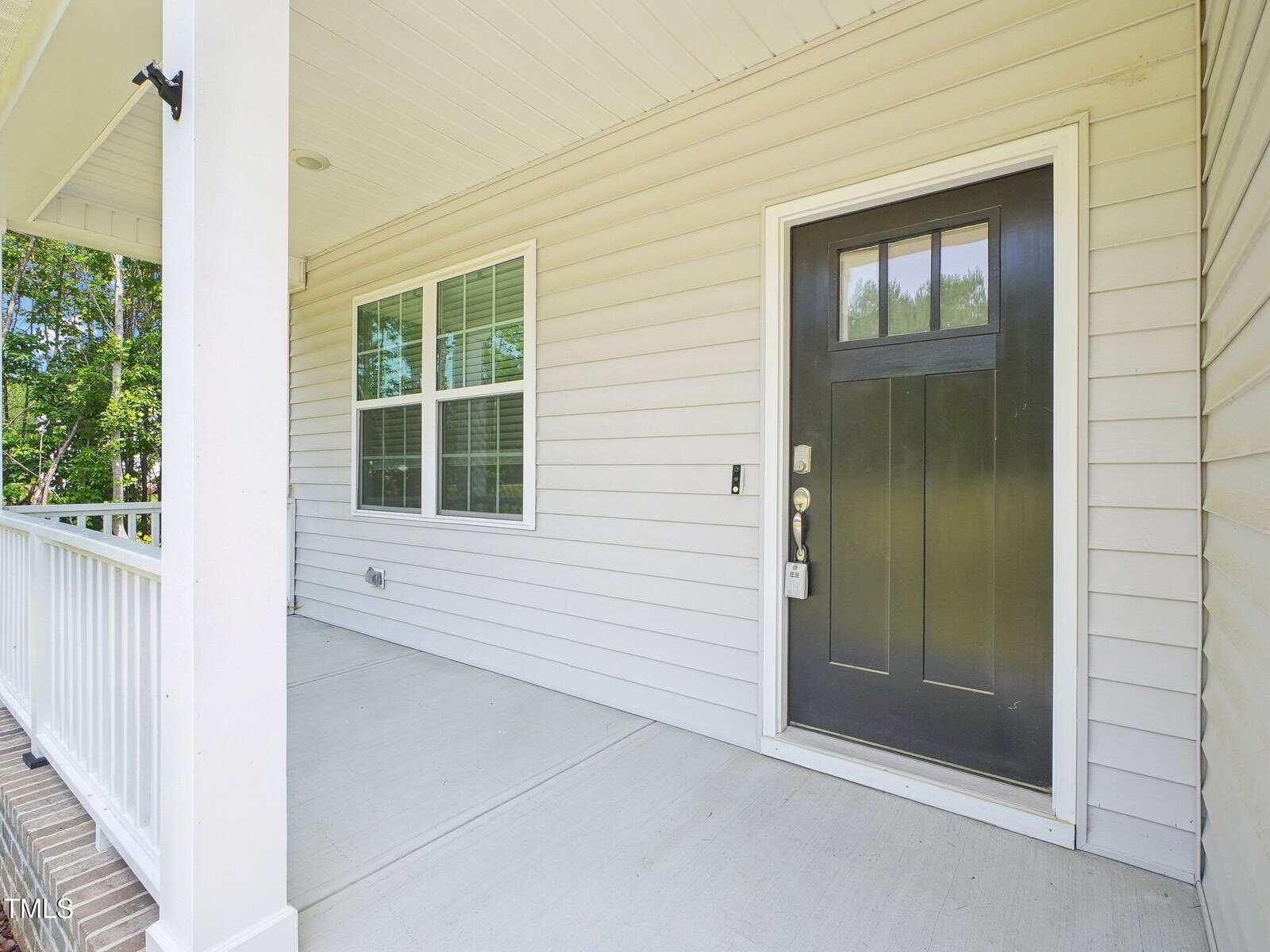 A covered front porch of a house with beige siding, a black front door with glass panels, white railing, and two large windows. The porch has a clean, light-colored concrete floor. Trees are visible in the background.