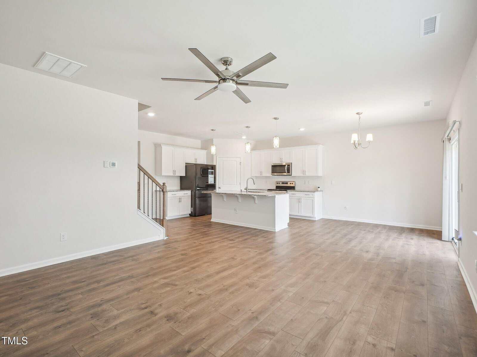 Spacious open-plan kitchen and living area with wood flooring, white cabinets, stainless steel appliances, ceiling fan, chandelier, staircase, and large windows letting in natural light.