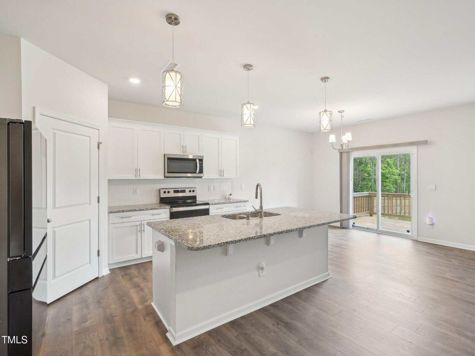 Modern kitchen with white cabinets, stainless steel appliances, granite island, pendant lighting, and hardwood floors. Sliding glass doors open to a deck with a view of greenery outside.