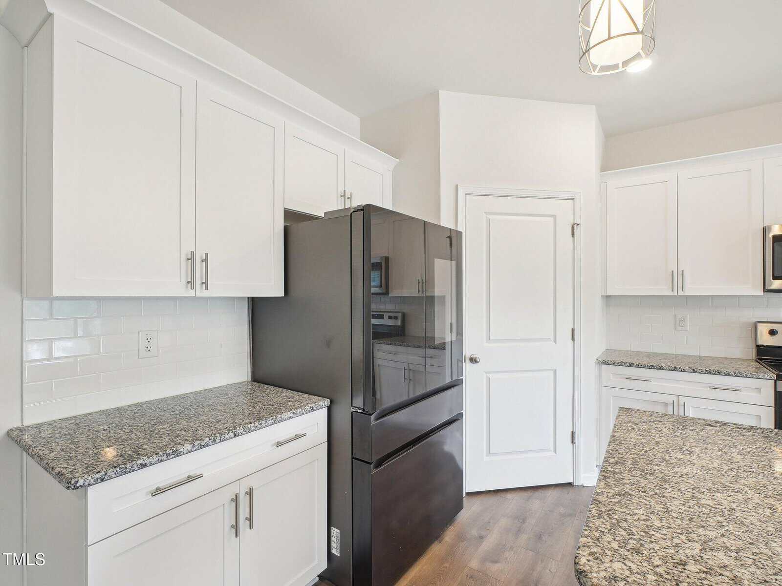 Modern kitchen with white cabinets, granite countertops, a black refrigerator, stainless steel microwave, white subway tile backsplash, and wood-look flooring. A light fixture hangs from the ceiling. Door and electrical outlet visible.