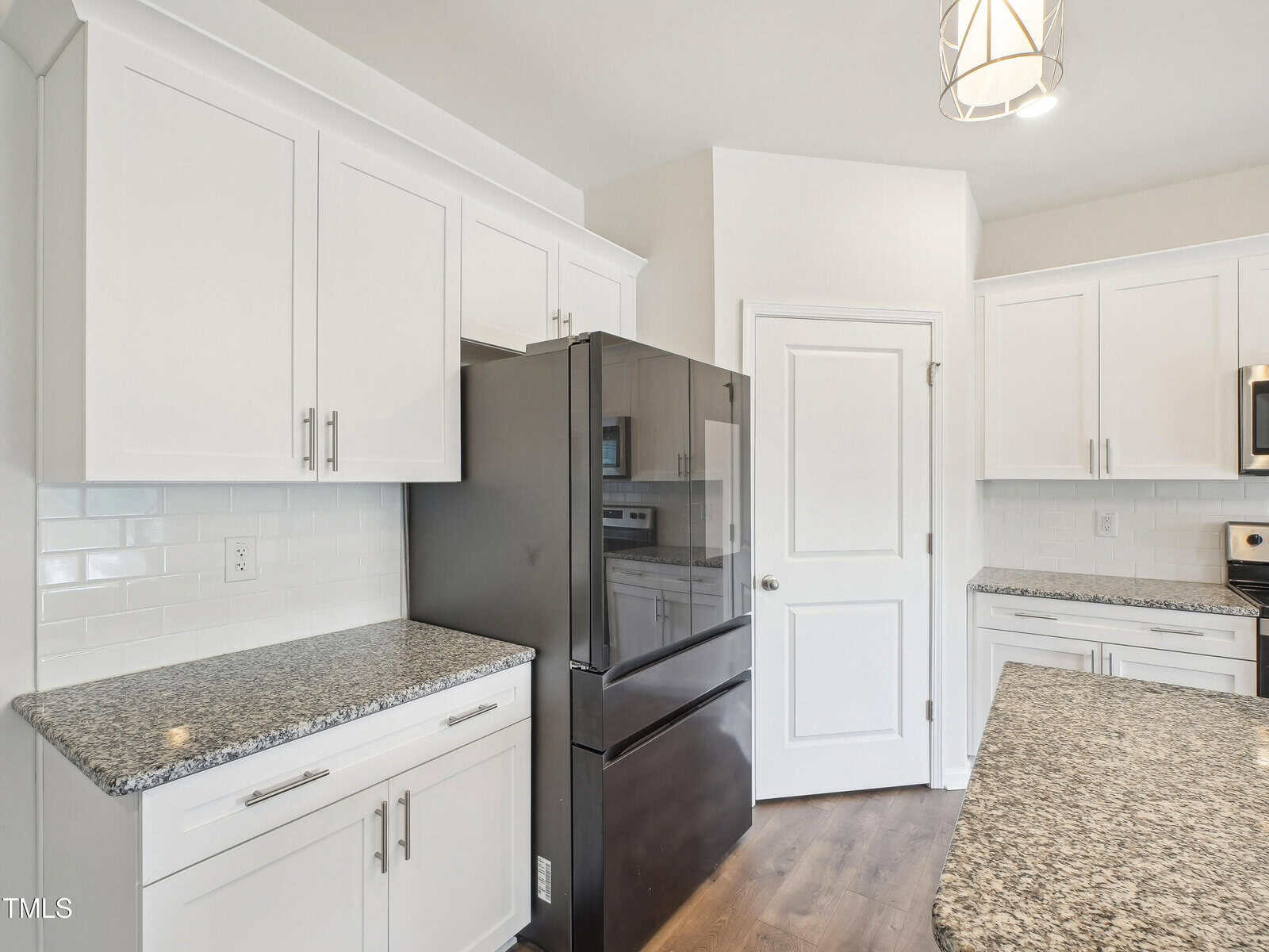 A modern kitchen with white cabinets, granite countertops, a black refrigerator, a white door, and wood flooring. A hanging light fixture is visible on the ceiling.