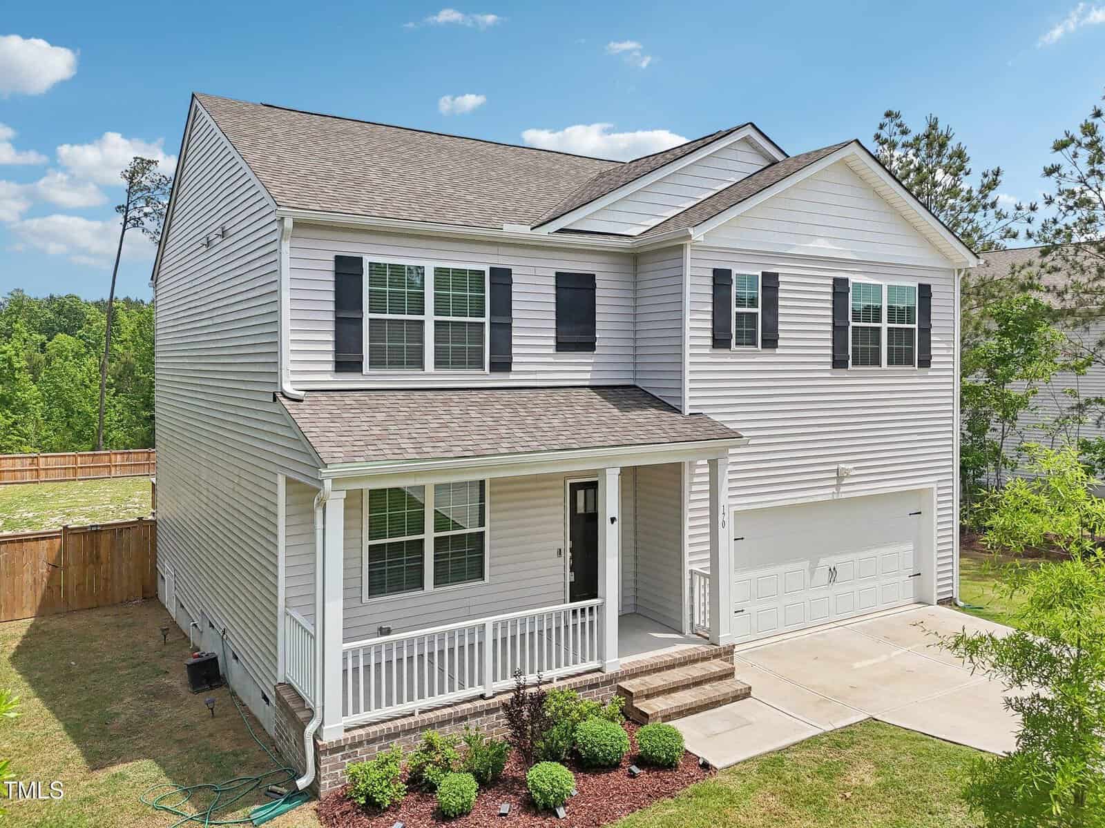 Two-story white house with black shutters, a covered front porch, attached two-car garage, and neatly landscaped front yard. The home sits on a lawn with a wooden fence and trees in the background under a blue sky.