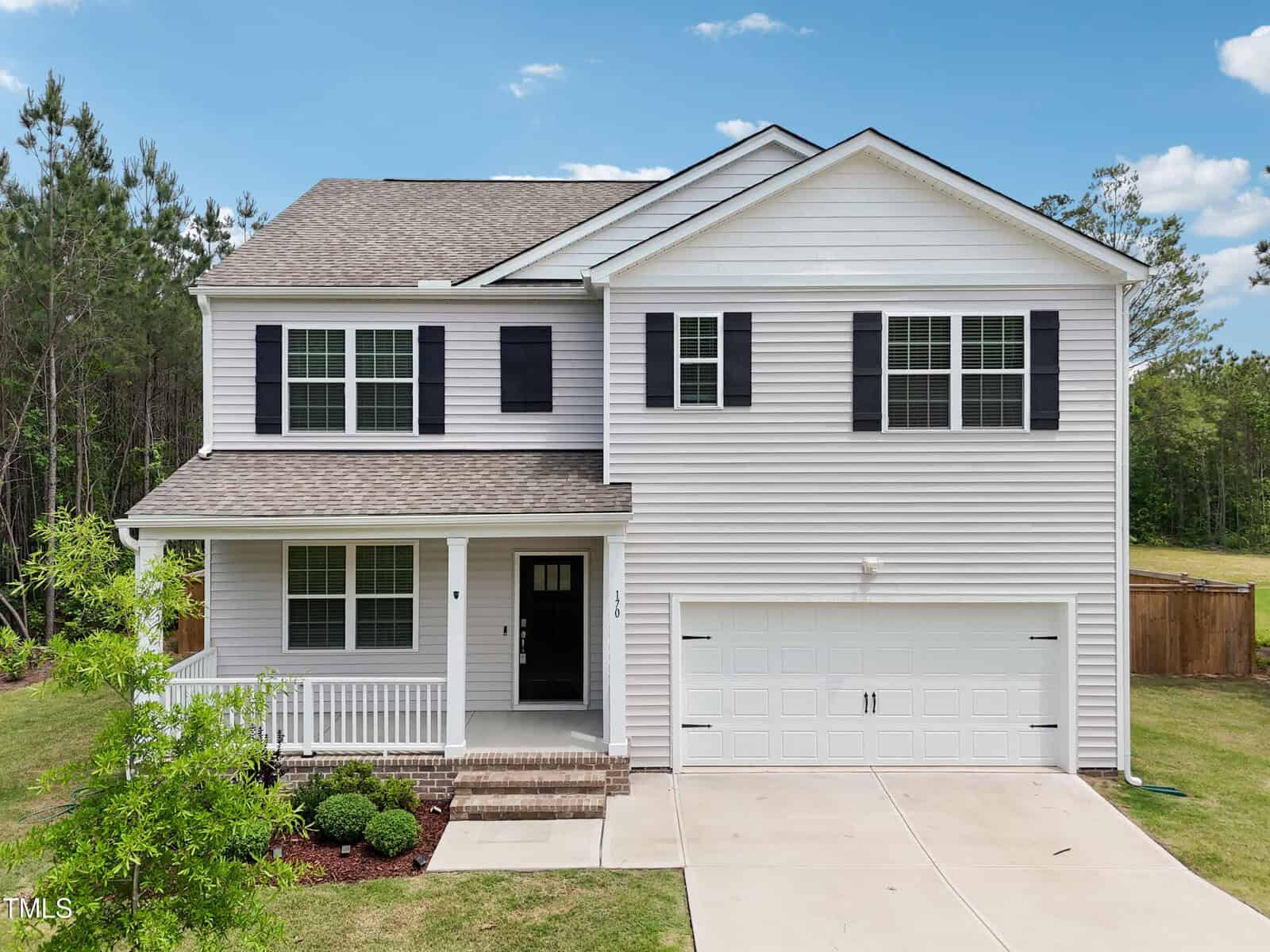 A two-story white house with black shutters, a covered front porch, and an attached two-car garage. The home is surrounded by greenery with trees and a wooden fence in the background under a blue sky.