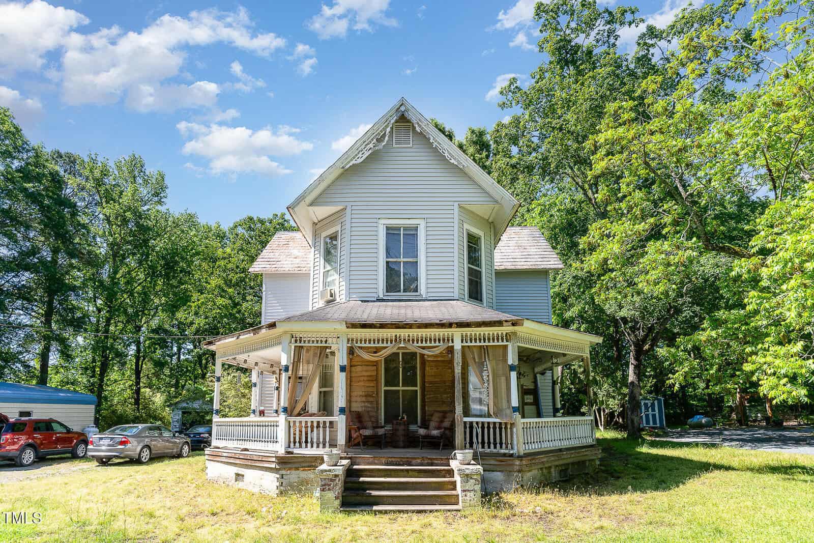 A two-story gray Victorian-style house at 110 E Church Street, Creedmoor, with a wraparound porch, ornate trim, and wood siding. Surrounded by trees and parked cars to the left, beneath a blue sky with scattered clouds.