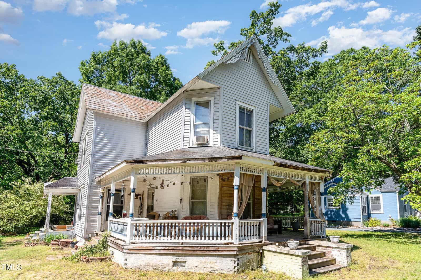 A two-story white wooden house with a covered wraparound porch, decorative trim, and surrounded by green trees, photographed on a sunny day with blue sky and scattered clouds.