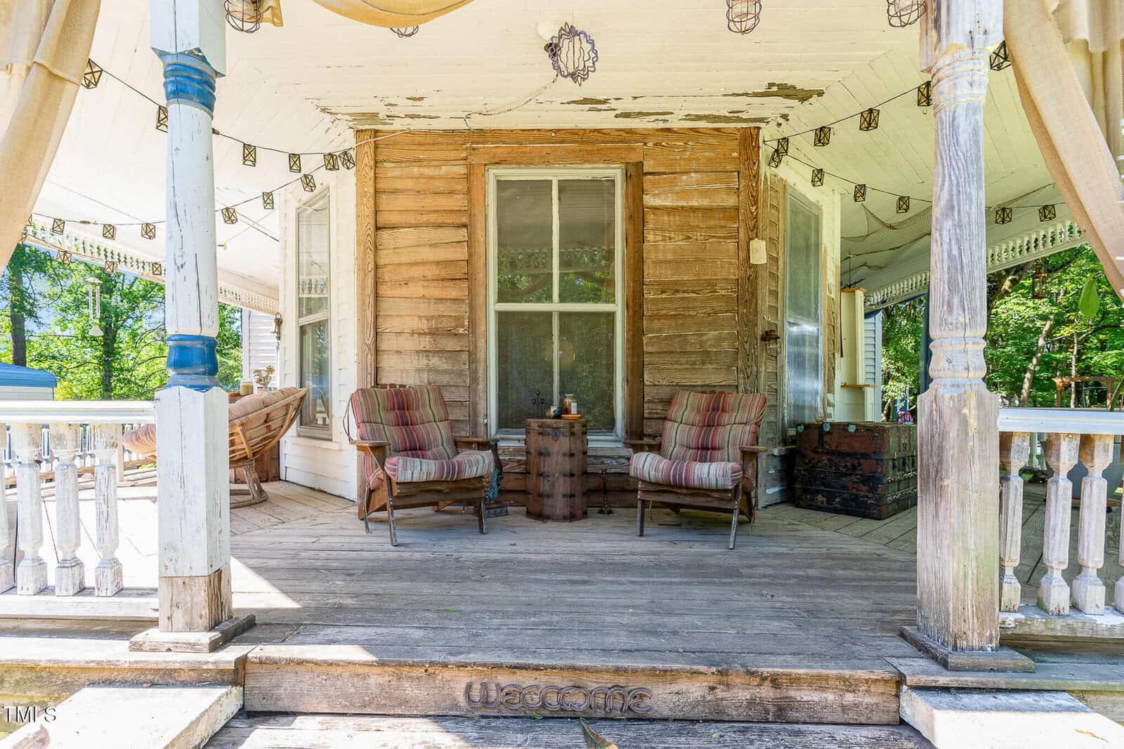 A weathered wooden porch with two striped cushioned chairs and a small table in front of a window. String lights hang above, and the word "Welcome" is on a mat at the base of the steps.
