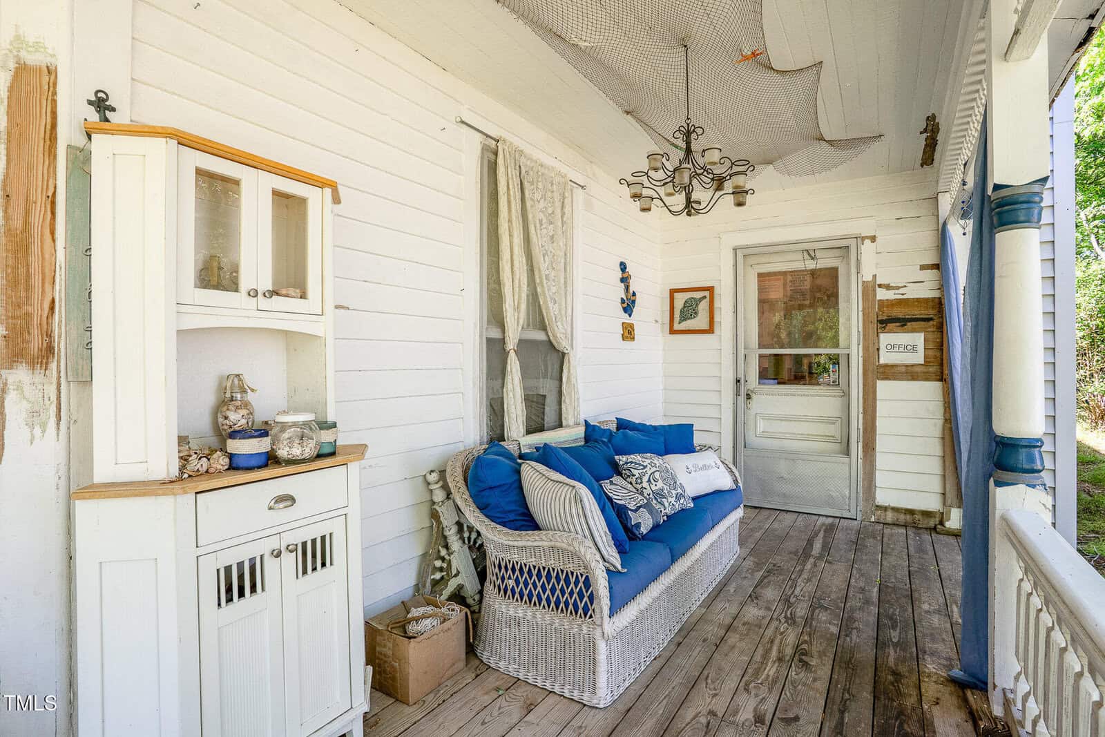A white wooden porch features a wicker sofa with blue and white cushions, a small white cabinet with glass jars, wall decor, a chandelier, and wood-plank flooring; sunlight shines through a doorway.