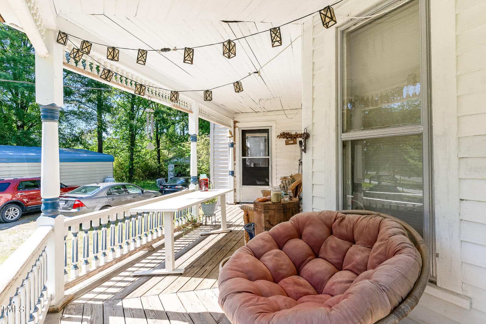 A covered wooden porch with string lights, a round cushioned chair, a rustic table, and a large window. Several cars are parked outside next to trees and a blue-roofed building.