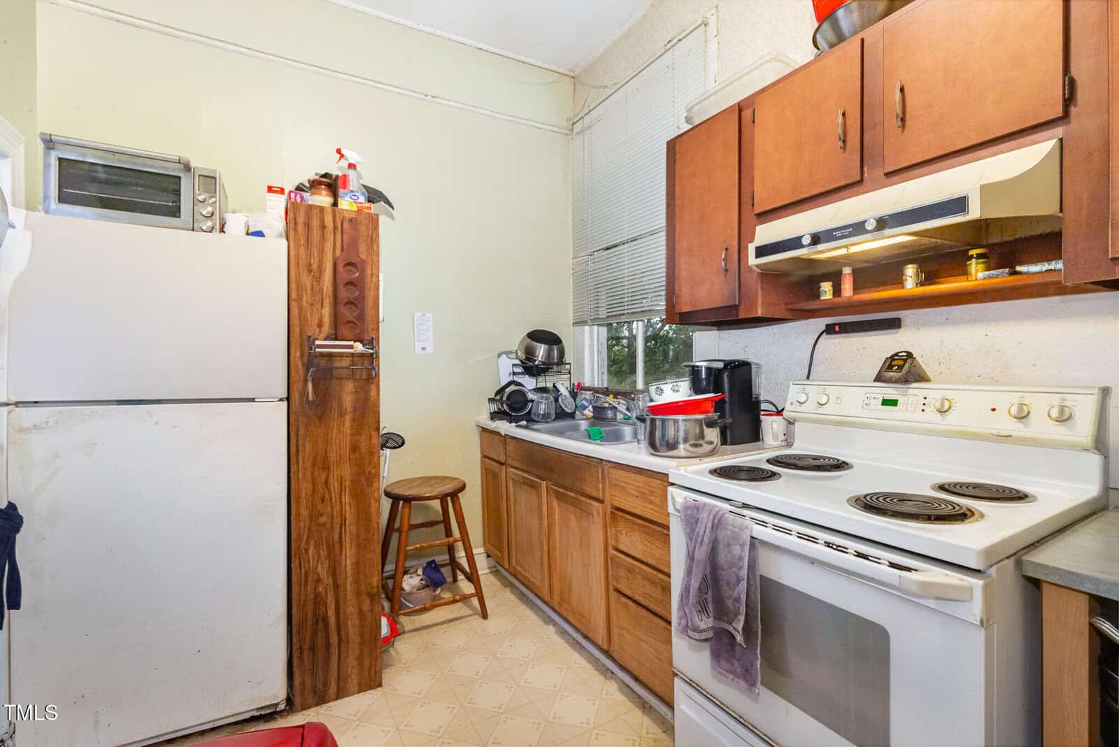 A small kitchen with a white fridge, microwave, and stove. There are wooden cabinets, various appliances on the counter, stacked dishes, and a stool beside the fridge. A towel hangs on the oven door.