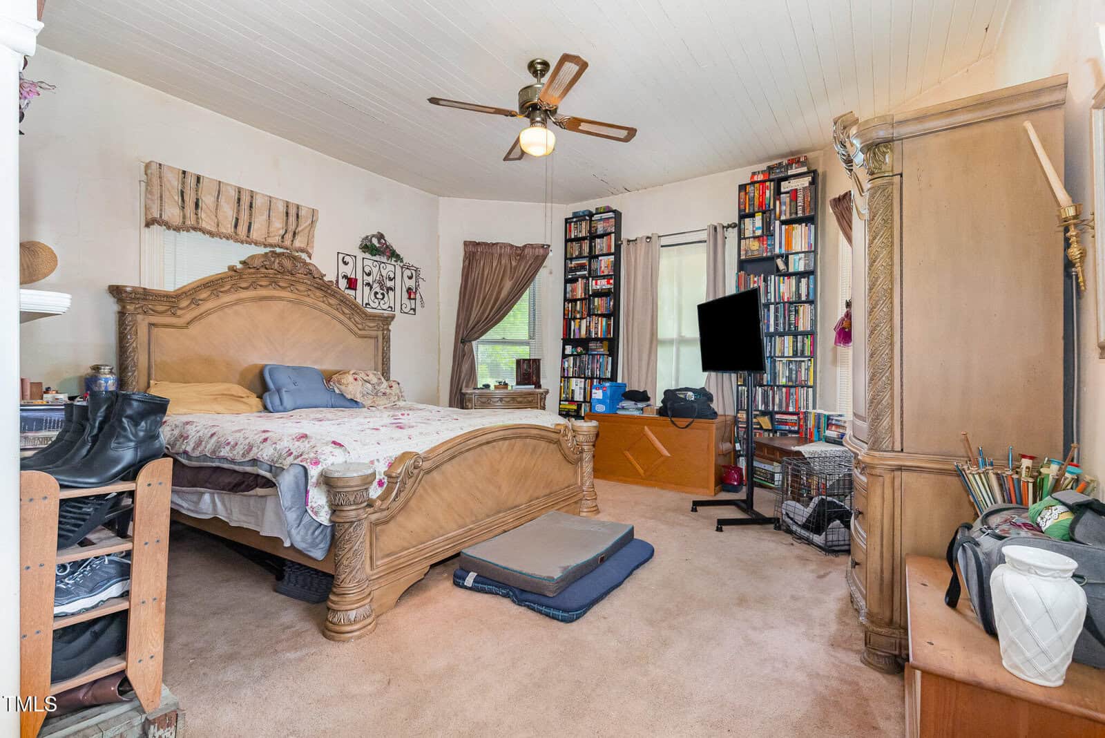 A bedroom with a large wooden bed, beige carpet, bookshelves filled with books and board games, a TV on a stand, shoe rack, and various personal items. A ceiling fan hangs from the white paneled ceiling.