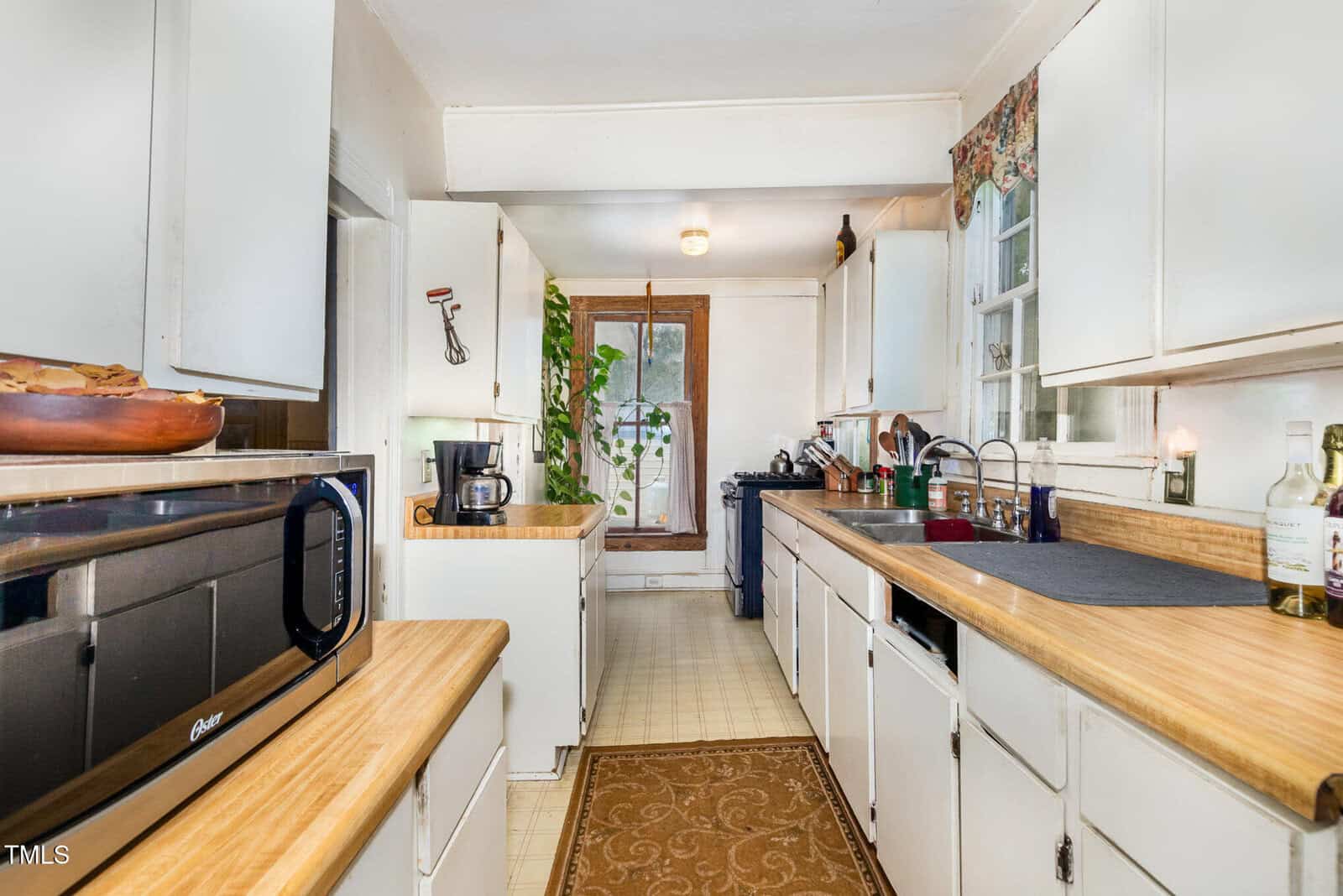 Narrow kitchen with white cabinets, wood countertops, and light-colored flooring. A rug runs along the floor, and there is a microwave, coffee maker, sink, and various small appliances on the counters. A door with glass panels is at the far end.