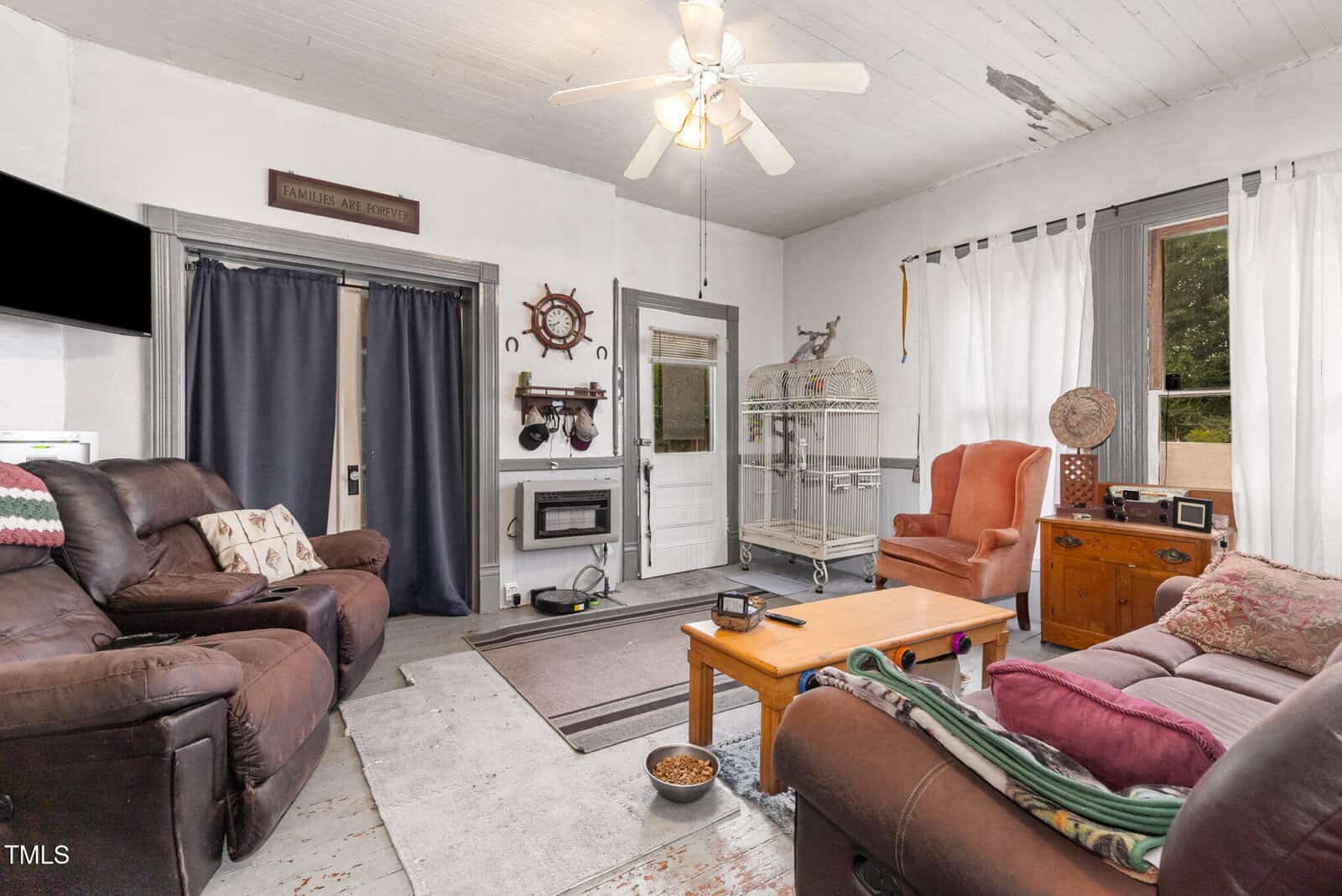 A living room with brown recliners, a green couch, a wooden coffee table, an orange armchair, a birdcage, wall-mounted TV, wood stove, large window with white curtains, and a ceiling fan.
