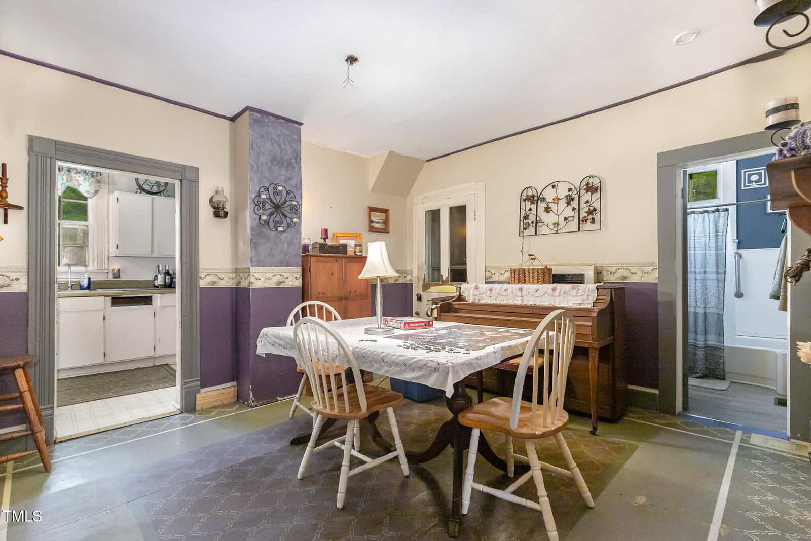 A dining area with a table, four chairs, a tablecloth, and a piano against the wall. Doors lead to a kitchen on the left and a bathroom on the right. The room has beige and purple walls with decorative accents.