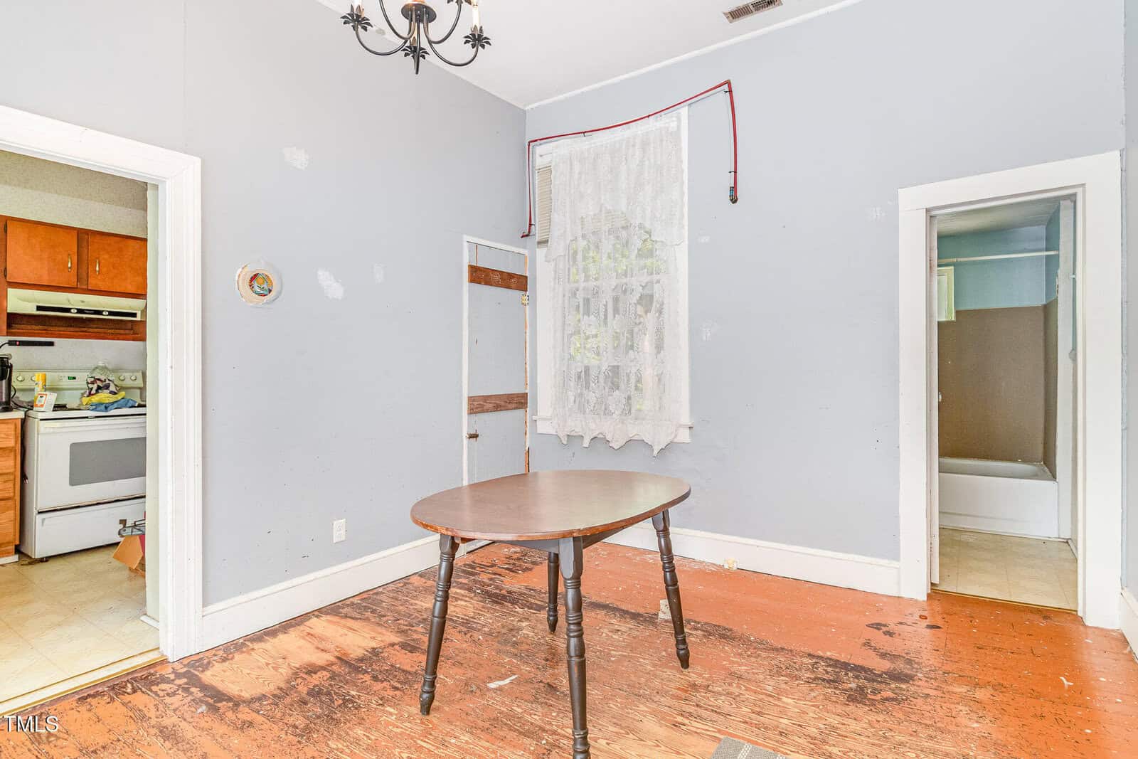 A small, sparsely furnished room with a round wooden table and worn, partially stripped floors. The walls are light blue with scuff marks. Doorways lead to a kitchen and a bathroom. A lace curtain covers the window.