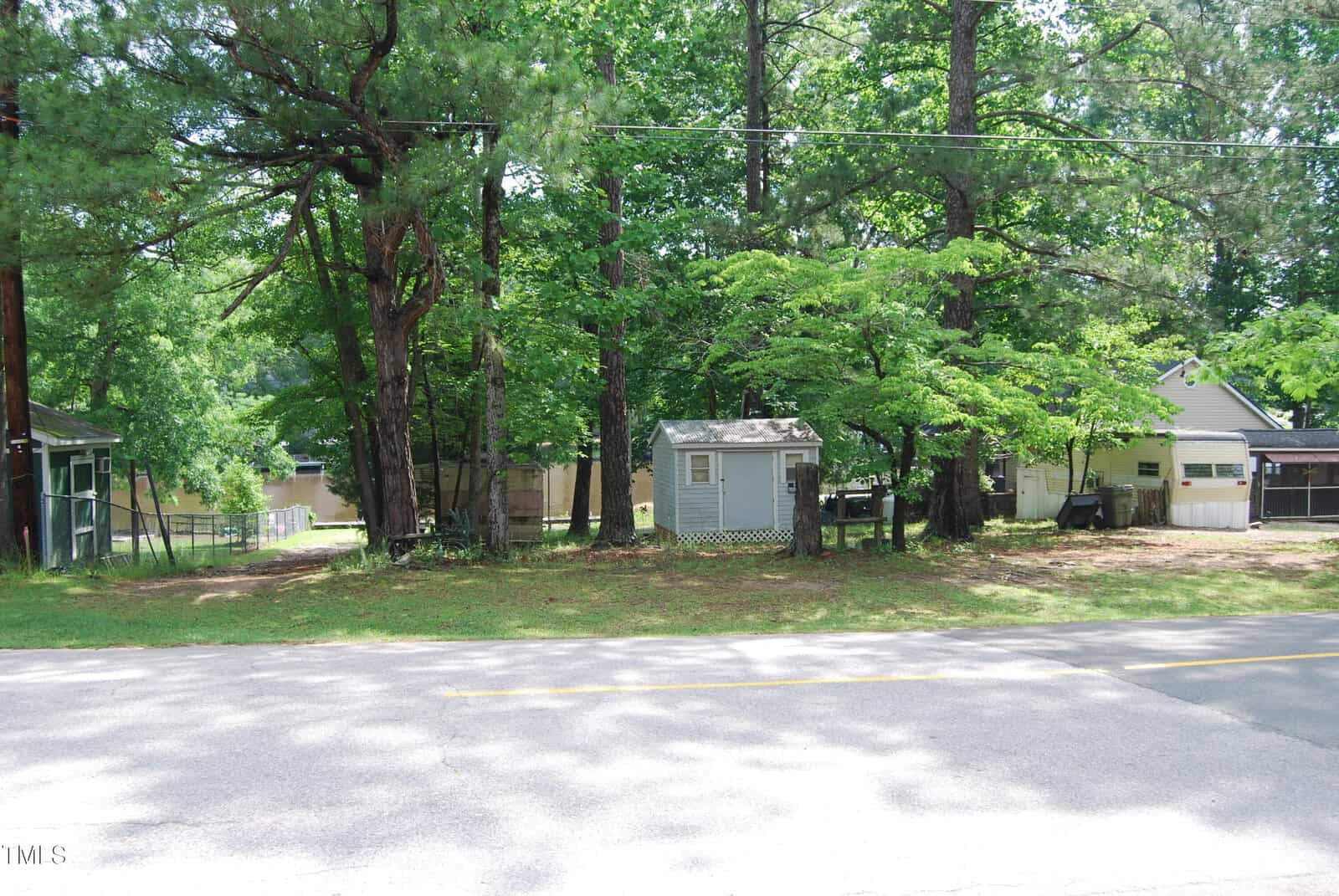 A small gray shed sits among tall trees and green foliage across a quiet street, with houses partially visible in the background. Sunlight filters through the leaves, casting dappled shadows on the ground.