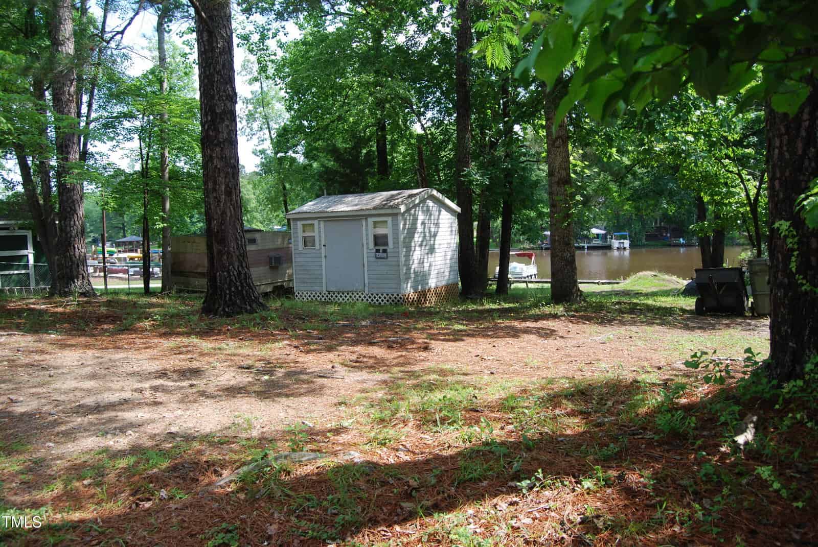 A small gray shed sits among tall trees on a patch of cleared ground near a river, with cabins and boats visible across the water in the background. Sunlight filters through the leaves, casting dappled shadows.