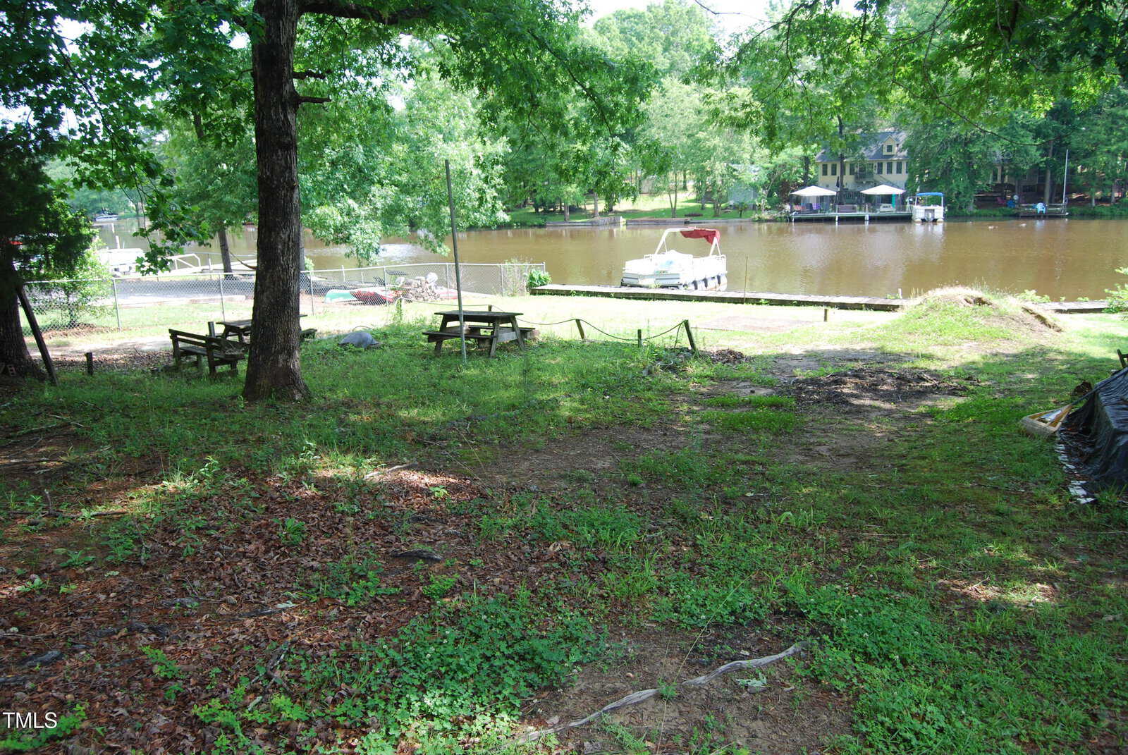 A grassy, partially shaded yard with benches and a tree overlooks a calm lake with docks, boats, and houses visible on the far shore. The scene is peaceful and surrounded by greenery.