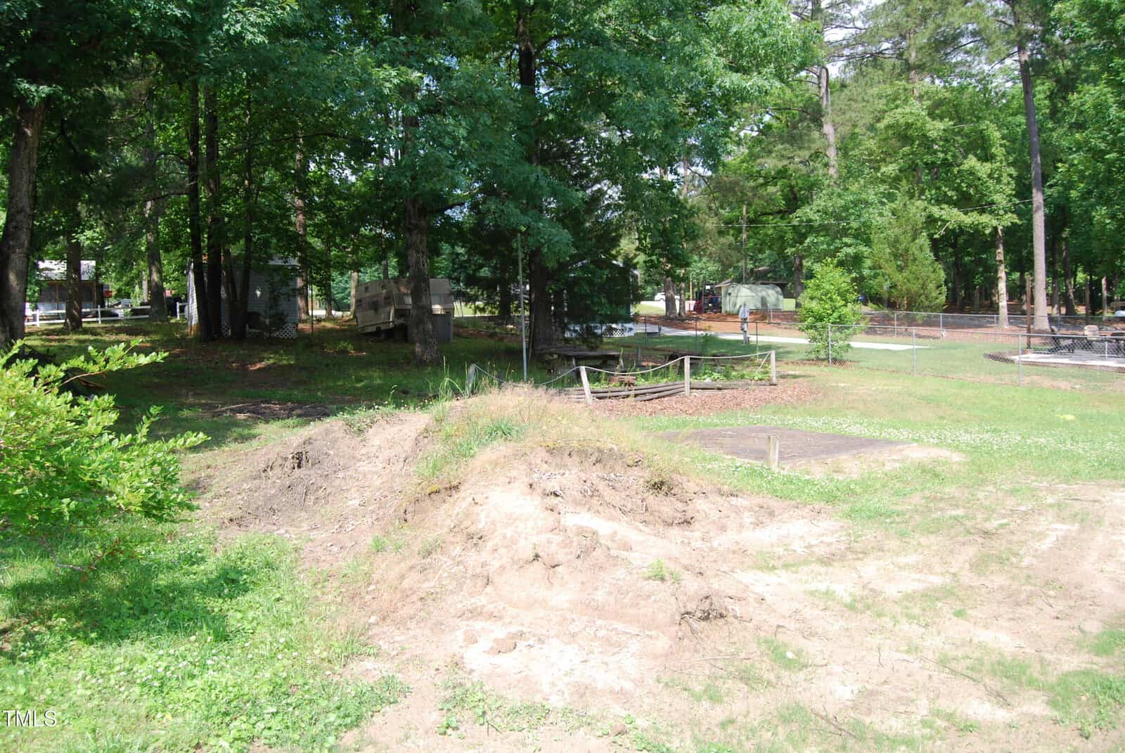 A sunlit, grassy yard with patches of bare dirt in the foreground, surrounded by tall trees. In the background, an RV, picnic tables, and fenced areas are visible among the trees.