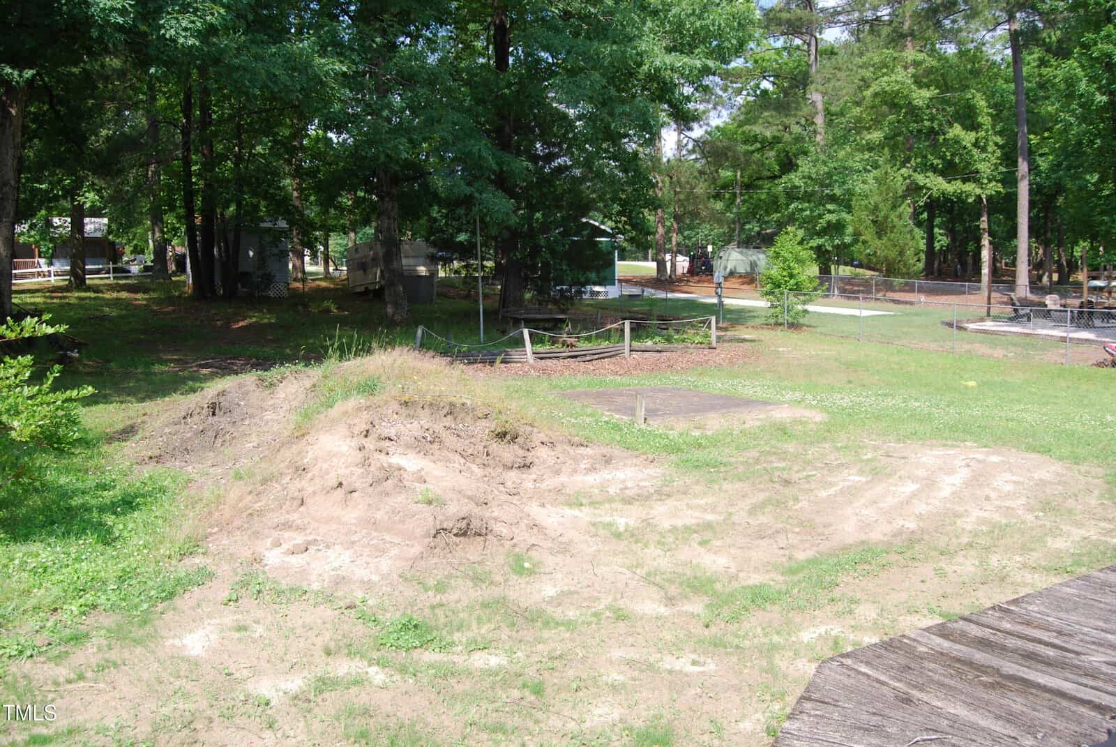 A partially cleared, grassy backyard with patches of dirt, small mounds of soil, and several trees. Fenced gardens and various structures are visible in the background, surrounded by greenery.