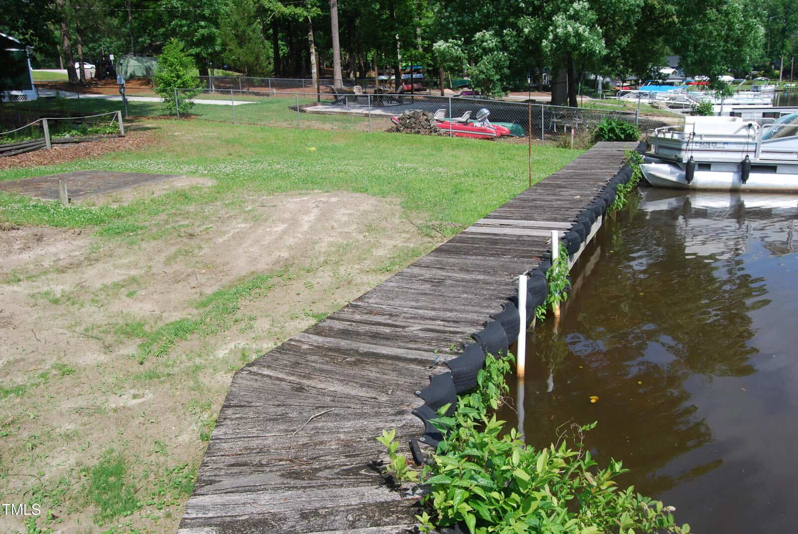 A wooden dock extends over a lake beside a grassy yard with sparse patches of dirt. Boats are tied nearby, and trees border the area in the background.