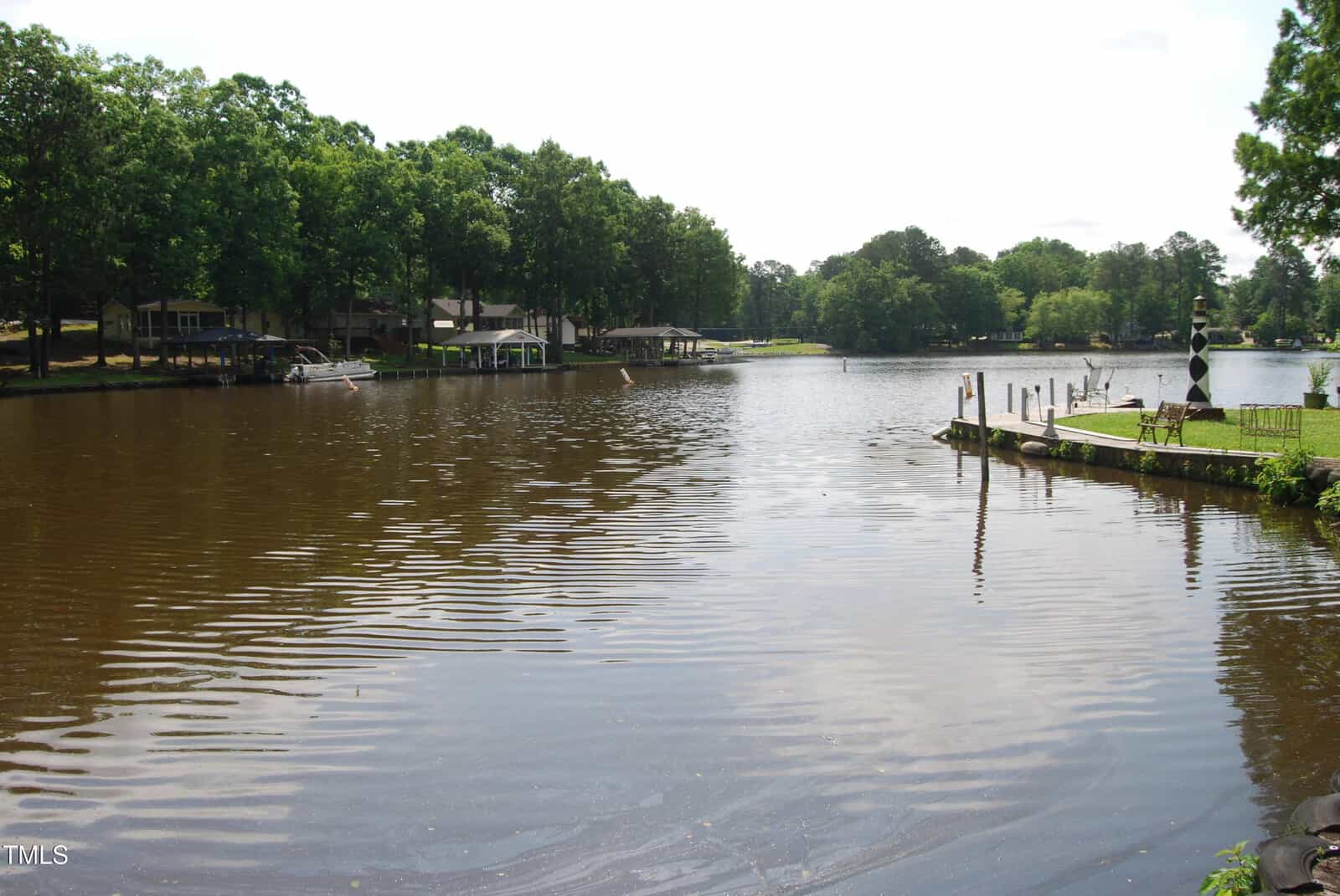 A wide, calm lake bordered by trees and houses with boat docks. The water is brownish, reflecting the greenery. A grassy area and wooden posts line the right edge of the image. The sky is mostly clear.